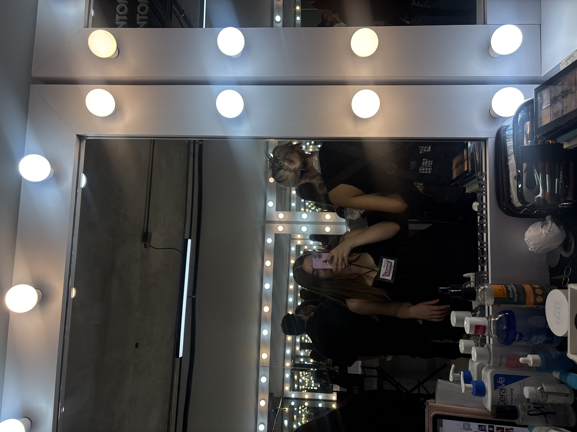 The image shows a mirror with a white frame and round light bulbs around it. Two women are visible in the reflection of the mirror. One woman is holding a phone up to her face. The other woman stands behind her and appears to be fixing her hair. There are several bottles of lotions, creams, and other cosmetic products in front of the mirror. Some of the brands visible are CeraVe, Nivea, and Alcohol Isopropilico. Part of a badge can also be seen on one of the women. Based on the presence of Spanish-language text on some of the bottles, and the OCR results showing the text "Mexico", the photograph was possibly taken in Mexico City, Mexico.