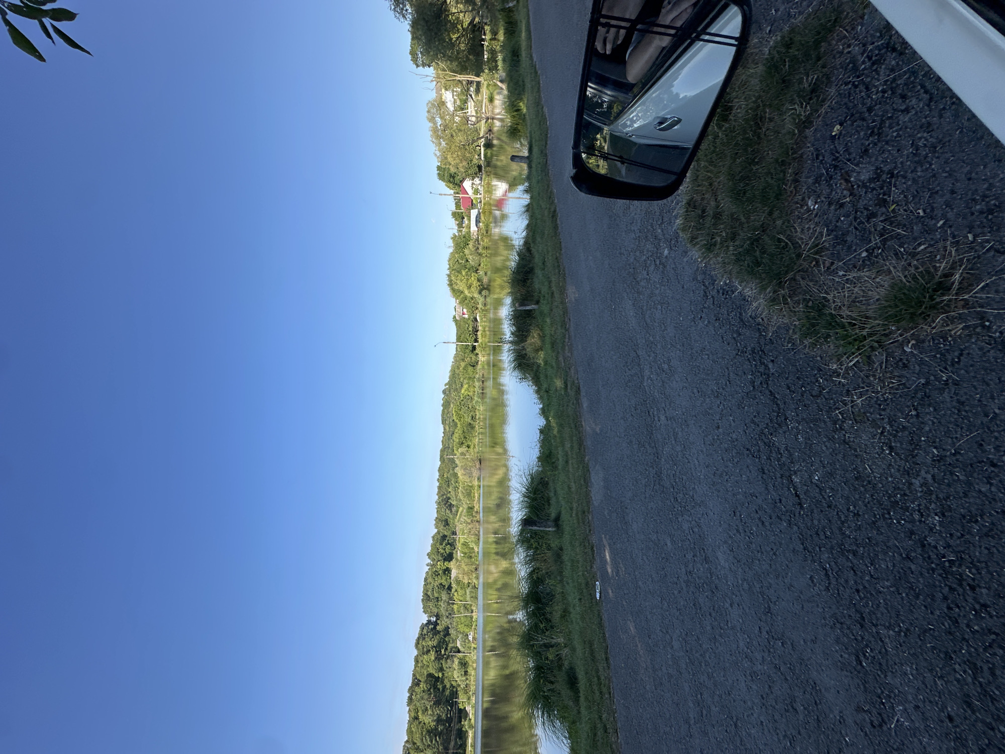 The image depicts a serene lake scene on a clear day, viewed from a vehicle. In the foreground, a rough asphalt road occupies the lower portion of the frame, with a patch of dry grass growing at its edge. To the right, a car's side mirror reflects the interior of the vehicle, showing a person's arm and shoulder. Beyond the road lies the lake, its calm surface mirroring the lush green trees and blue sky. The far bank of the lake is lined with a dense forest. Several houses with red roofs are visible nestled among the trees on the right side of the lake, suggesting a settlement near the water. The overall impression is one of tranquility and natural beauty, possibly on the outskirts of a town or village. It is plausible this scene is located in Makiv, Ukraine, a region known for its picturesque landscapes.