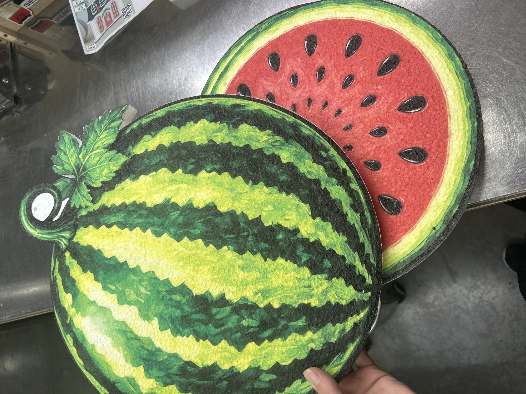 A close-up overhead shot shows a hand holding a decorative cutout of a watermelon. The cutout is a two-part design, with the rind and top of the watermelon on the left, and a slice of the watermelon, showing the red flesh and black seeds, on the right. The watermelon cutout is placed on a metallic surface, possibly a table or counter. In the background, there are glimpses of a wooden crate and some paper packaging, suggesting this might be a retail or storage environment. The lighting is bright, casting slight shadows. The image is taken from a high angle, looking down at the watermelon cutouts. The scene is devoid of any specific landmarks or context that would place it in Ternopil, Ukraine.