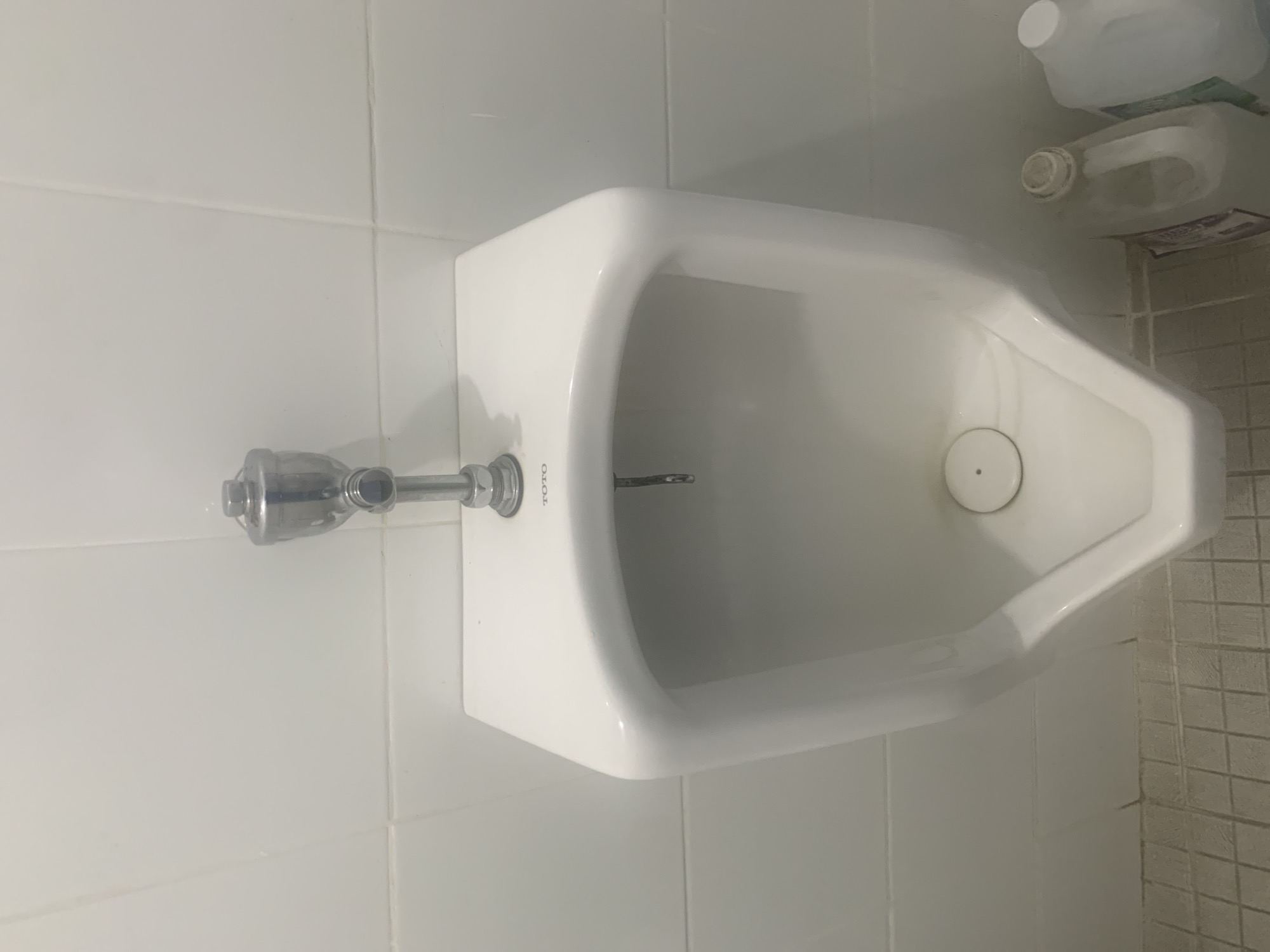 A white, wall-mounted urinal with a chrome flush valve is centrally located in the frame, against a white tiled wall. A TOTO logo is visible on the urinal. The urinal has a V-shaped basin with a drain cover at the bottom. To the right of the urinal, two large translucent plastic jugs are visible on the tiled floor. The jug on the left has a white cap and a white handle, and the jug on the right also has a white cap. The floor is covered in beige square tiles. The scene appears to be in a public restroom in Palembang, Indonesia.