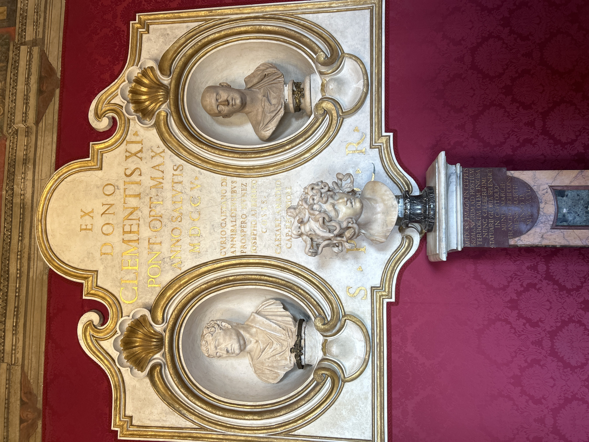 The image displays a wall-mounted marble plaque with three marble busts, set against a richly patterned deep red damask wall covering. The scene is located within the Capitoline Museums, specifically the Palazzo dei Conservatori, in Rome, Italy.
The central feature is a large, ornate plaque composed of light-colored marble or stucco with extensive gilded accents. It includes a central inscription area and two oval niches. The top portion of the plaque bears a gilded Latin inscription reading: "EX DONO CLEMENTIS XI PONT OPT MAX ANNO SALUTIS MDCCV" (From the gift of Clement XI, Pontifex Optimus Maximus, in the year of salvation 1705). Below this are further gilded Latin texts, partially visible. Below the two upper busts, separated by the lower central bust, are the gilded letters "S.P." on the left and "R." on the right, signifying "Senātus Populusque Rōmānus."
Within the left oval niche, a marble bust of a male figure with short, curly hair and wearing a toga is presented, looking slightly to the left. In the right oval niche, a marble bust of a bald male figure wearing a toga is presented, looking directly forward. Both busts are classical Roman in style and rest on small, ornate gilded pedestals.
Centered directly beneath the main plaque, a third, smaller marble bust of Medusa is displayed. This bust depicts a head with hair stylized as intertwining snakes and an intense facial expression. It rests on a dark, ornate pedestal, which in turn sits atop a larger, rectangular marble plinth. The plinth for the Medusa bust features its own Latin inscription, detailing its historical context and dating to "MDCCXXXI" (1731).
The wall covering is a deep red, possibly silk, damask fabric featuring an intricate floral and foliage pattern. Above the main plaque, a glimpse of an ornate gilded architectural cornice is visible. The entire display is uniformly illuminated.