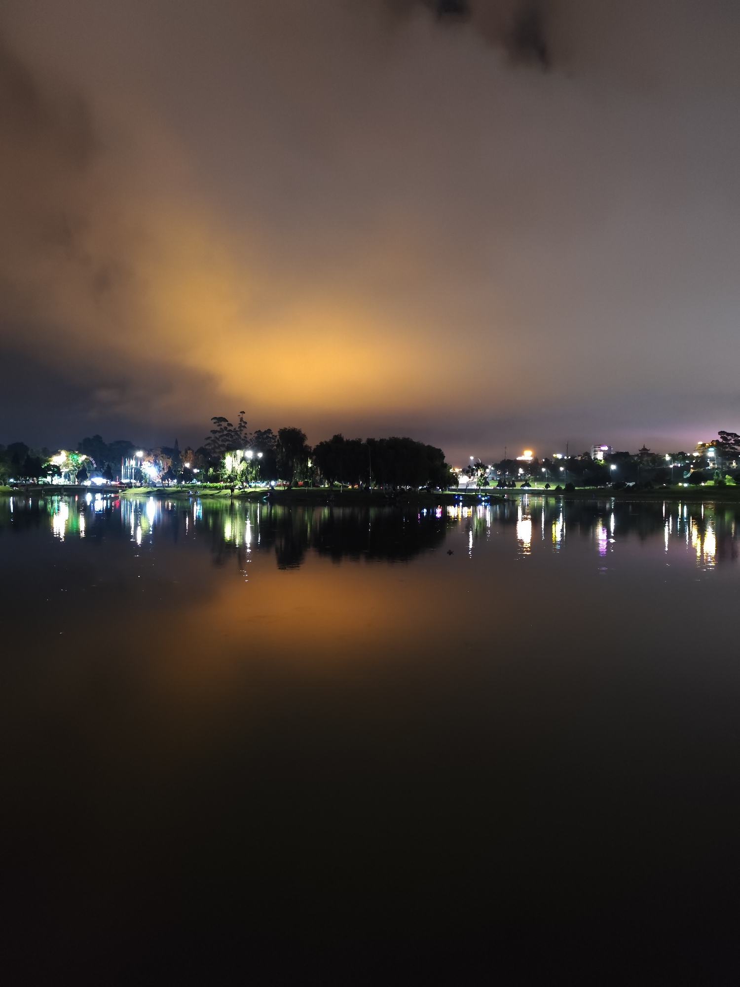 The image shows a night view of Ho Xuan Huong Lake in Da Lat, Vietnam. The lake's surface is mostly dark, reflecting the ambient light and creating shimmering reflections of the illuminated cityscape on the opposite shore. The horizon line is defined by trees and buildings, dotted with lights that create distinct reflections. Above, the sky is covered with dark clouds, and diffused orange lighting illuminates the sky.