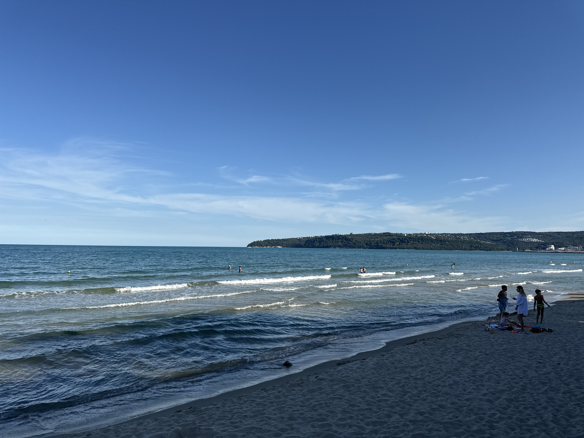 The image depicts a sandy beach with calm, blue-green ocean waves gently lapping at the shore under a clear, bright blue sky with wispy white clouds. A distant, tree-covered headland with scattered buildings is visible on the horizon. On the beach, a family group of four, silhouetted by the sun, stands near beach towels and other scattered items, appearing to be engaged in conversation. In the water, several individuals are swimming or wading, some further out and some closer to the shore. A small yellow buoy is visible in the distance, and a light-colored building structure with a tower is present on the far right coastline. The overall atmosphere is one of a tranquil summer day at the beach.