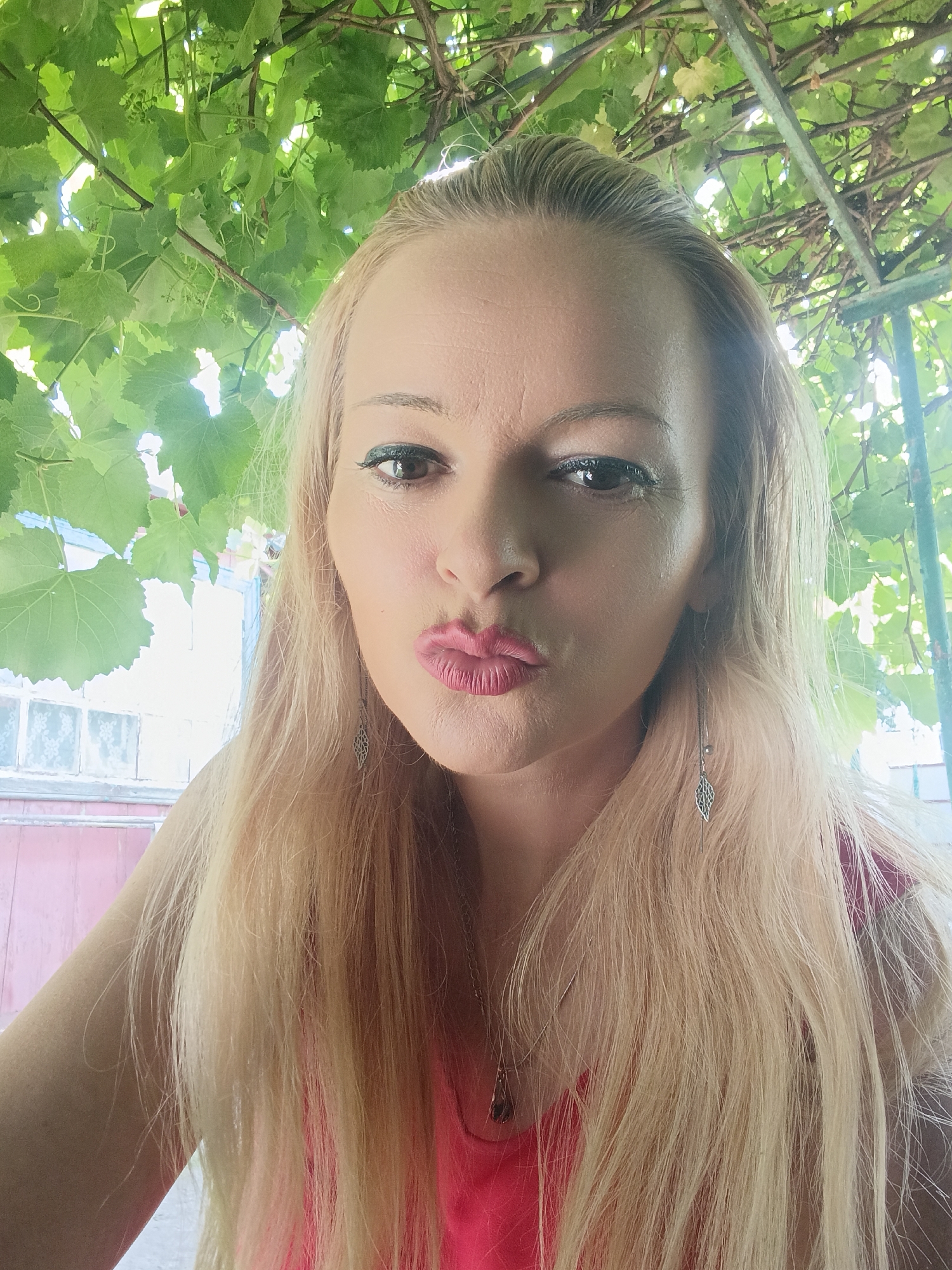 A blonde woman with fair skin and fair skin is taking a selfie outdoors. She is wearing pink clothing and makeup, with glitter eyeshadow and dark eyeliner. She has a leaf-shaped earring in her visible ear. Her lips are pursed, and she is looking directly at the camera. In the background, there is lush green foliage, suggesting she is under a tree or a vine-covered structure. A portion of a pink building and a window can be seen to her left, and to her right, a green metal structure is visible. The lighting is natural and bright, indicating it is daytime.