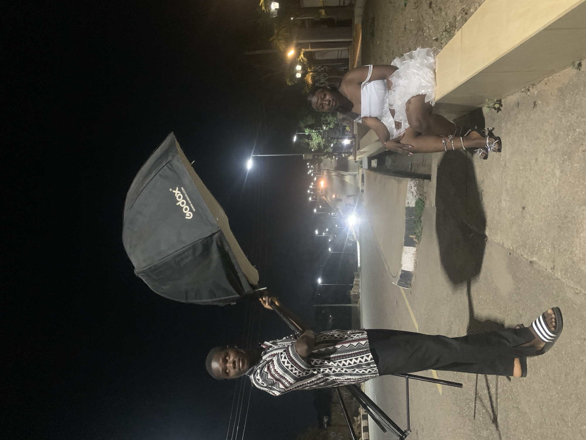 The image captures a nighttime outdoor photoshoot taking place in Jos, Nigeria. A woman in a white dress and heeled sandals is posing while sitting on a ledge or wall. She is the subject of the photograph. A man, likely the photographer or assistant, stands on the left side of the frame, holding a large, black, umbrella-like photography lighting modifier with the name "Godox" visible on its surface. He is dressed in a patterned shirt and dark pants and sandals. The background features a street with streetlights and some greenery. The location appears to be urban. The action consists of a staged photoshoot in an outdoor setting.