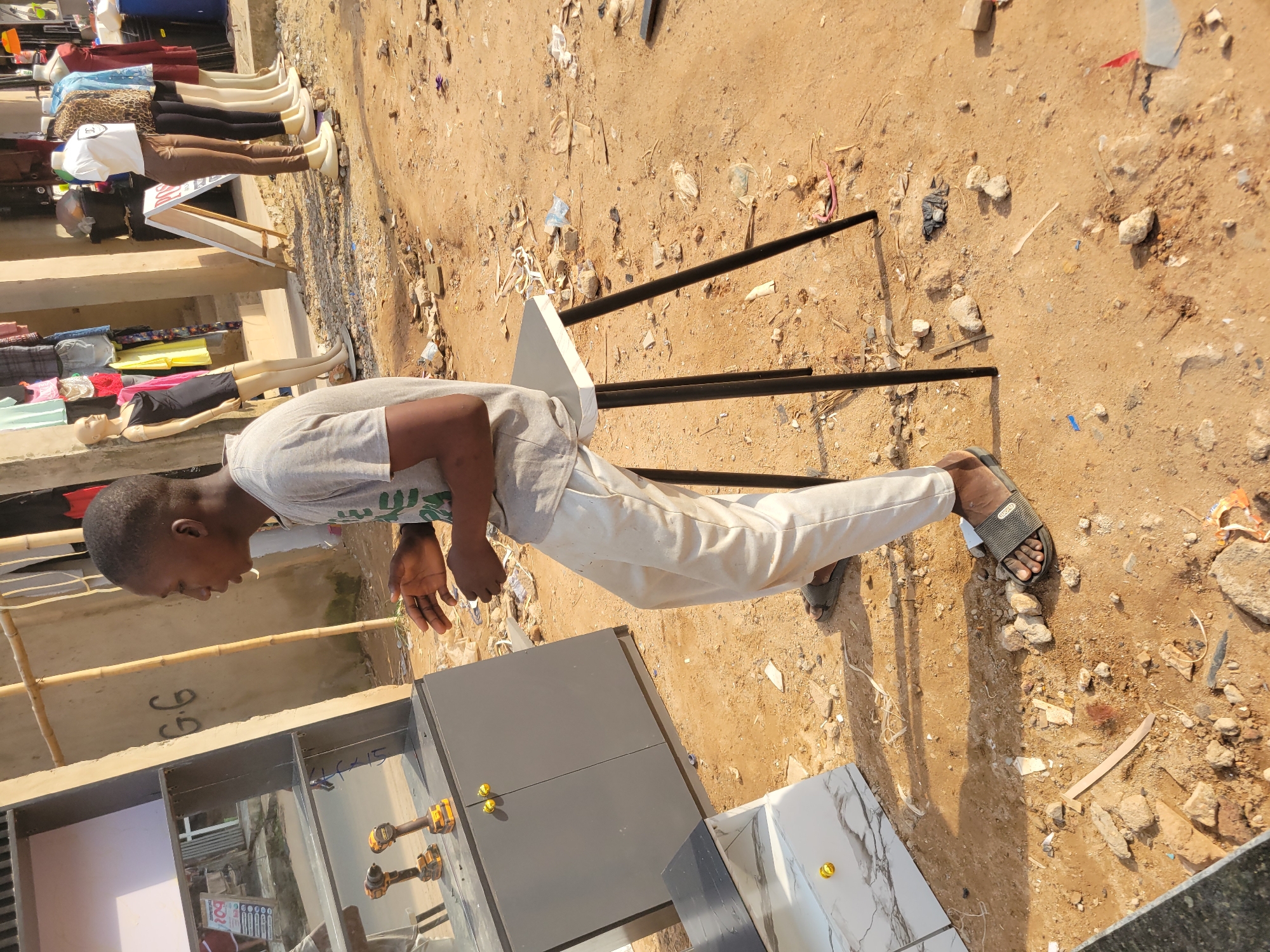 A young African male, appearing to be in his late teens or early twenties, is seated on a black-legged stool in an outdoor market. He wears a grey t-shirt and white trousers, with grey sandals. His head is turned to the right, and his mouth is slightly open as if speaking. In front of him, to the left, is a dark grey cabinet with two gold knobs and a mirrored surface on top. Two yellow power drills are visible on the cabinet's surface. Further to the left, a white and grey marble-patterned cabinet with a gold knob is partially visible. In the background, mannequins dressed in various clothing items are displayed along a building. The ground is dusty and littered with debris. The scene is consistent with an outdoor market setting in Egbagu, Nigeria.