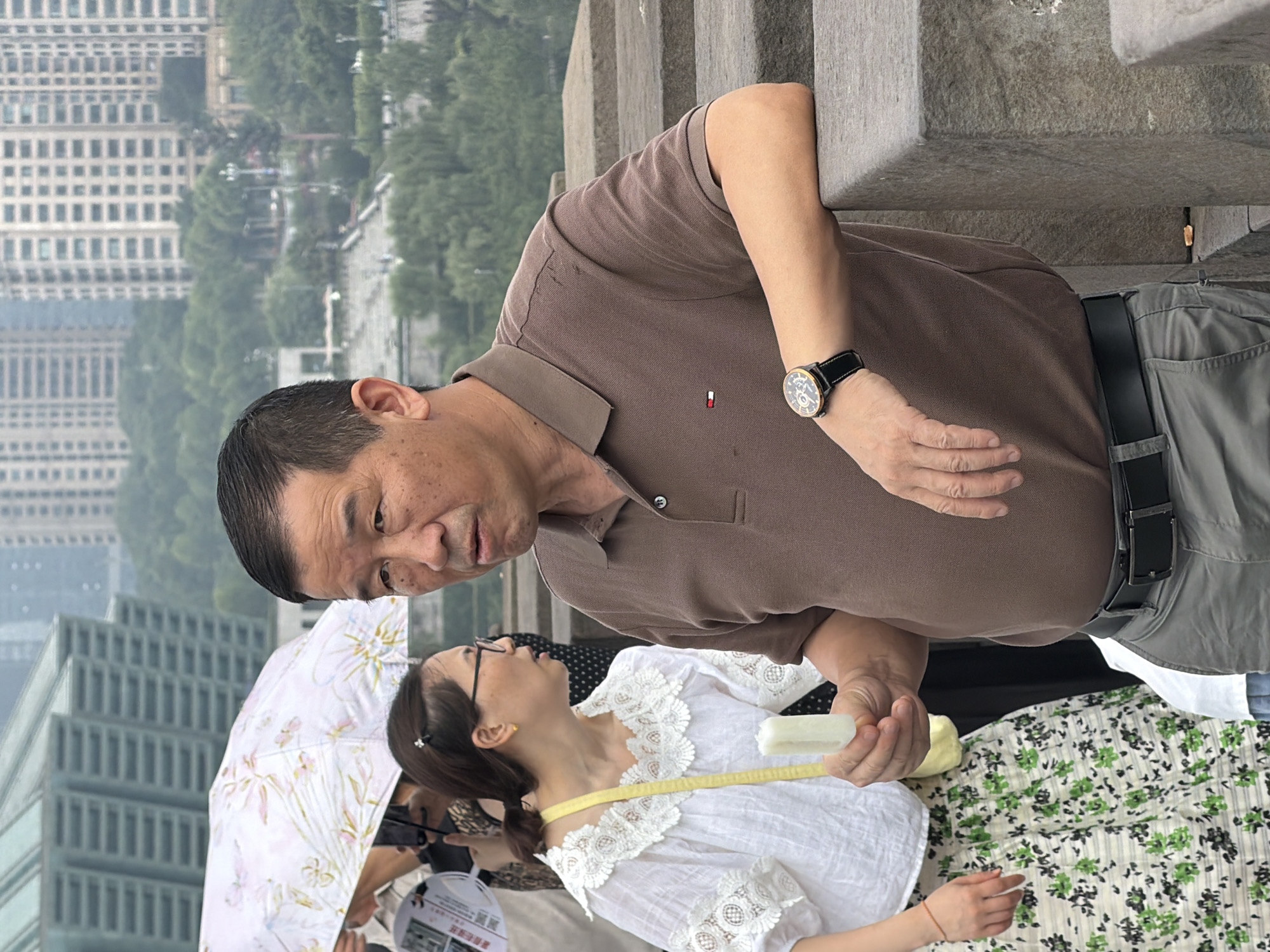 An outdoor, medium shot captures an East Asian man in his 50s or 60s wearing a brown polo shirt and grey trousers, leaning on a stone ledge. He is holding a white popsicle in his right hand. His left arm is bent, resting on the ledge, and he wears a black-strapped watch with a gold casing on his left wrist. Behind him, a woman in a white lace-trimmed top and a floral skirt is visible, also holding a popsicle. In the background, a patterned umbrella shields others, and a blurred cityscape with tall buildings and lush greenery suggests an urban setting. The overall scene appears to be in Yuzhong District, China.
