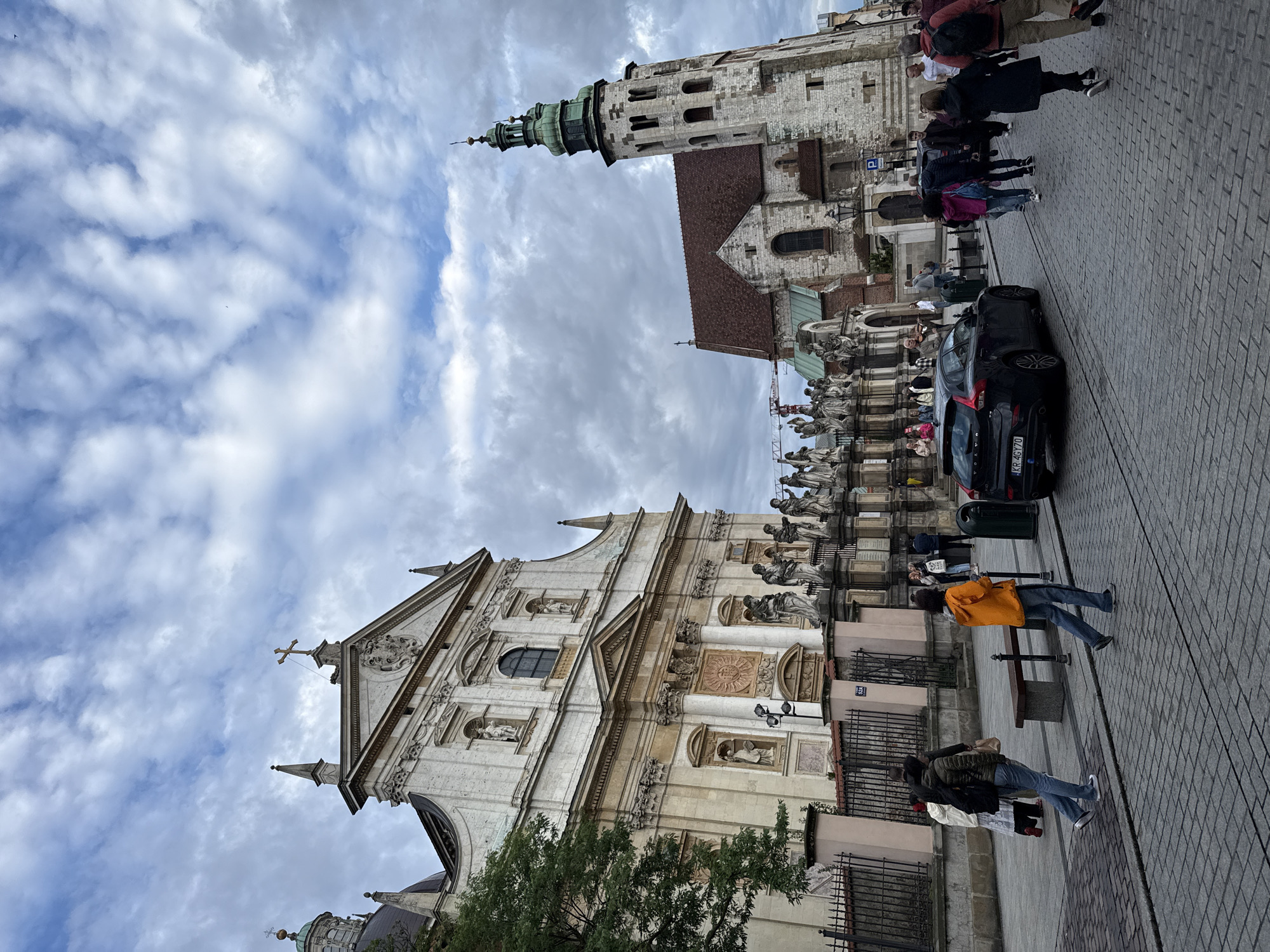 This is a street-level, medium-angle, full shot of the Main Market Square in Krakow, Poland, on a cloudy day. The iconic St. Mary's Basilica is visible on the right, a tall, stone structure with a green spire. To the left is the Lesser Basilica of St. Francis of Assisi, an ornate building with a white facade adorned with statues and columns. In the foreground, people are walking on a cobblestone street, and a black Volvo car is parked near the center. A street musician plays the violin beside the car. The sky is filled with dramatic, grey clouds, suggesting an overcast or approaching storm.