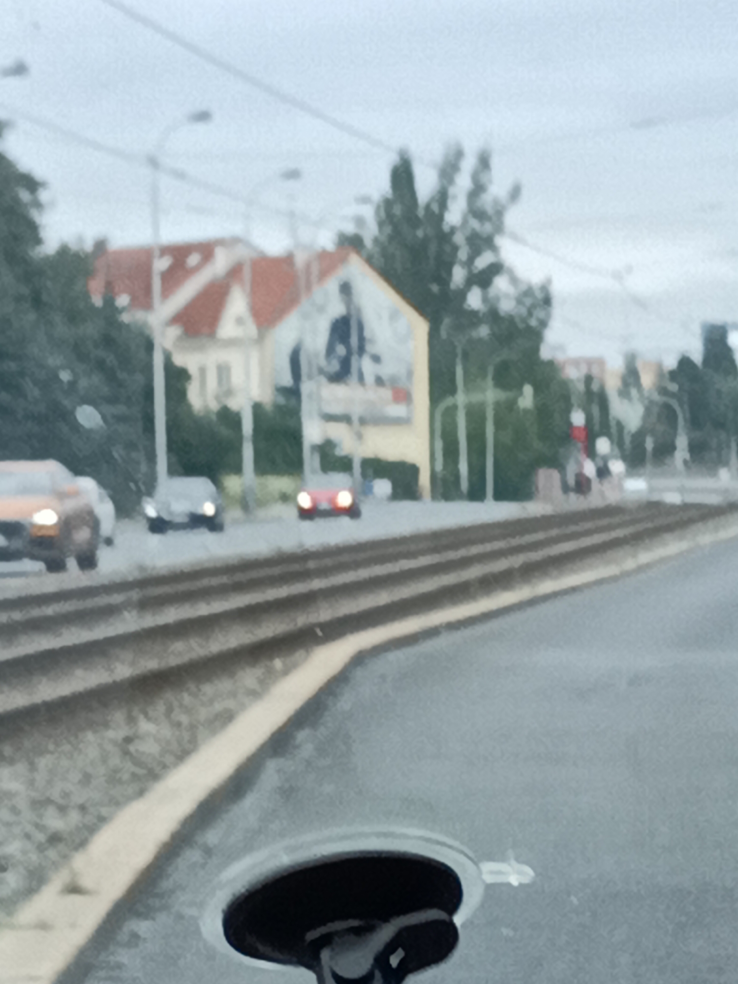 This is a photograph taken from inside a vehicle, likely with a phone camera, given the blurriness and the presence of a suction cup mount in the foreground. The image depicts a street scene with tram tracks running alongside the road.
In the background, several cars are visible on the road, including an orange car on the left, a dark car in the middle, and a red car on the right. Behind the cars, there are buildings with red-tiled roofs, some of which appear to be residential. One building has a large advertisement or mural on its side, showing a dark silhouette of a person. Trees and streetlights are also present, contributing to the urban setting. The sky is overcast, suggesting a cloudy or possibly rainy day. The overall lighting is diffused.
The image appears to have been taken during the daytime, but the cloudy conditions create a subdued light. The presence of tram tracks suggests the location is a city with a public transportation system. The architectural style of the buildings could offer clues to the specific location, but without further information, it is difficult to pinpoint. The general atmosphere is that of a typical urban commute or street view. There is no discernible text in the image.