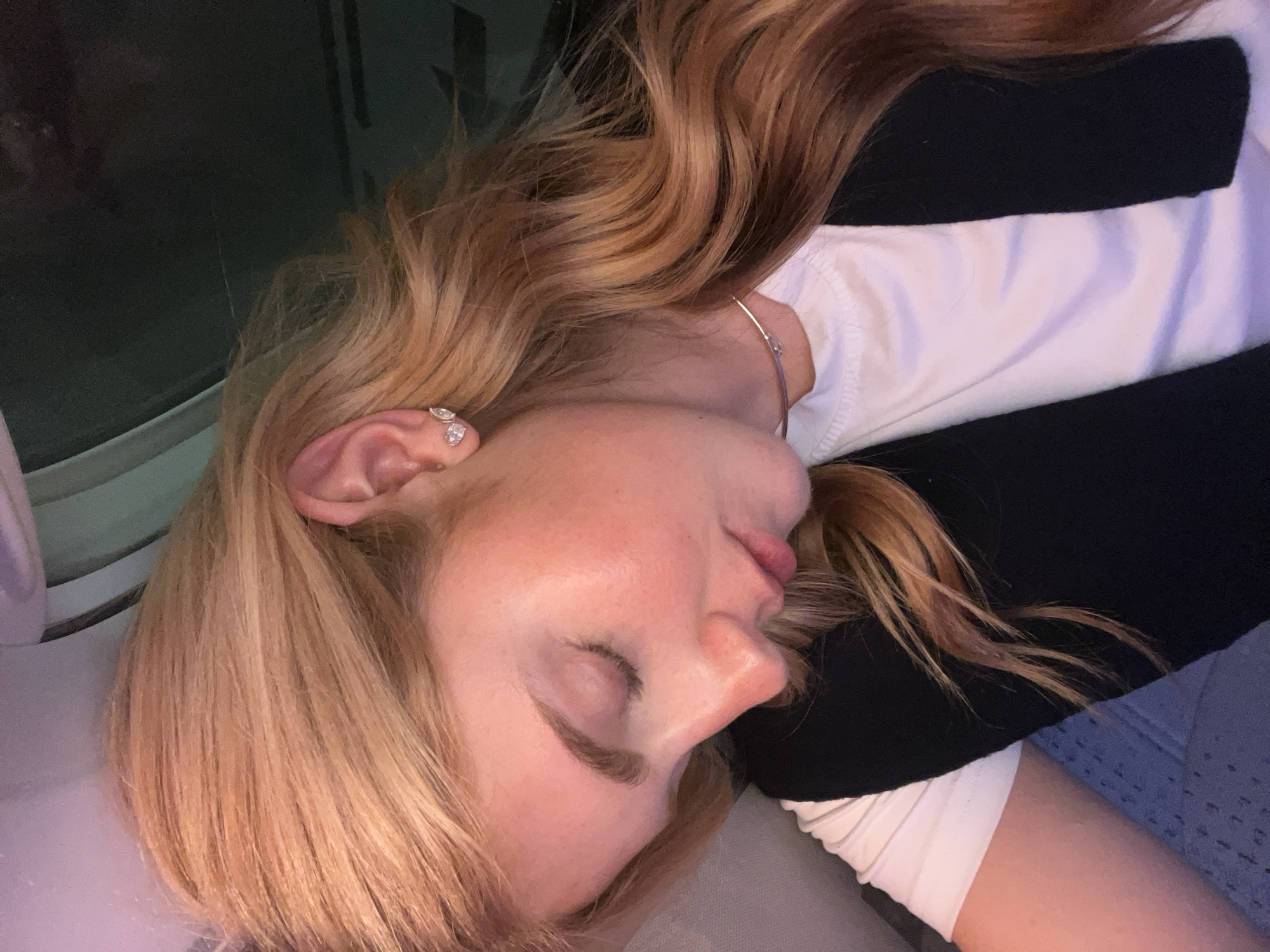 A person is lying down with their eyes closed, seemingly asleep. The person has fair skin and light blonde hair with waves. They are wearing a white shirt with a black top layered over it. The scene appears to be indoors, possibly on a plane, as there is a window visible in the upper left corner. The location is unspecified and cannot be determined as Kempton Park, South Africa.