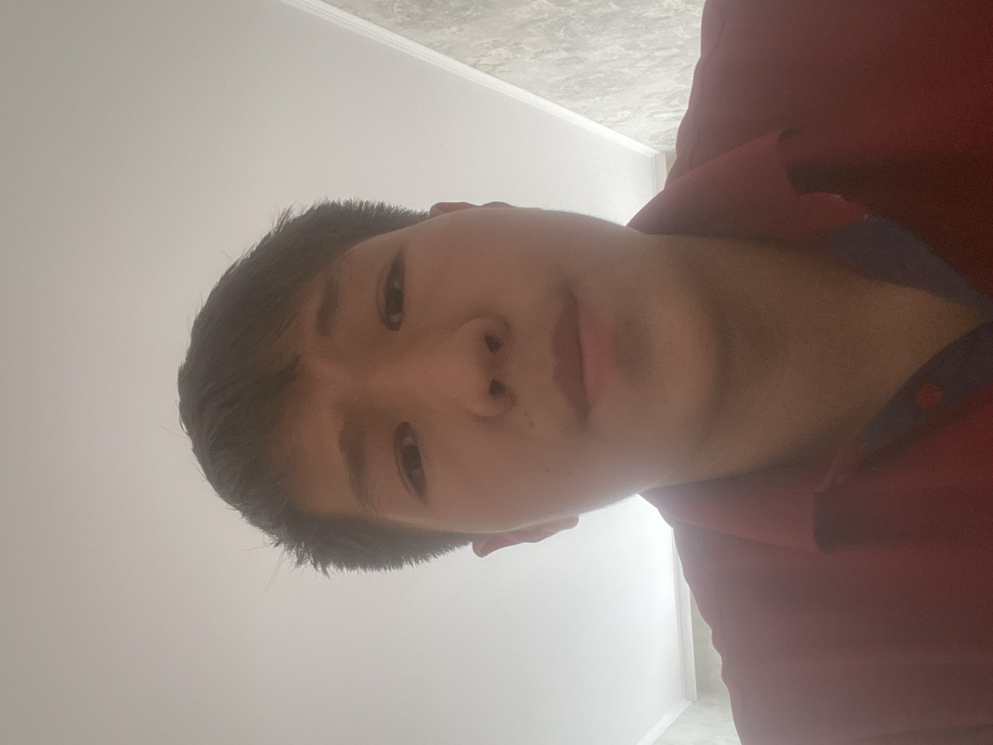 A young person with dark hair is looking directly at the camera in a selfie. They are wearing a red collared shirt. The background appears to be a ceiling with a textured wall to the right. The lighting suggests an indoor environment. The image is a close-up shot focusing on the upper body and face.