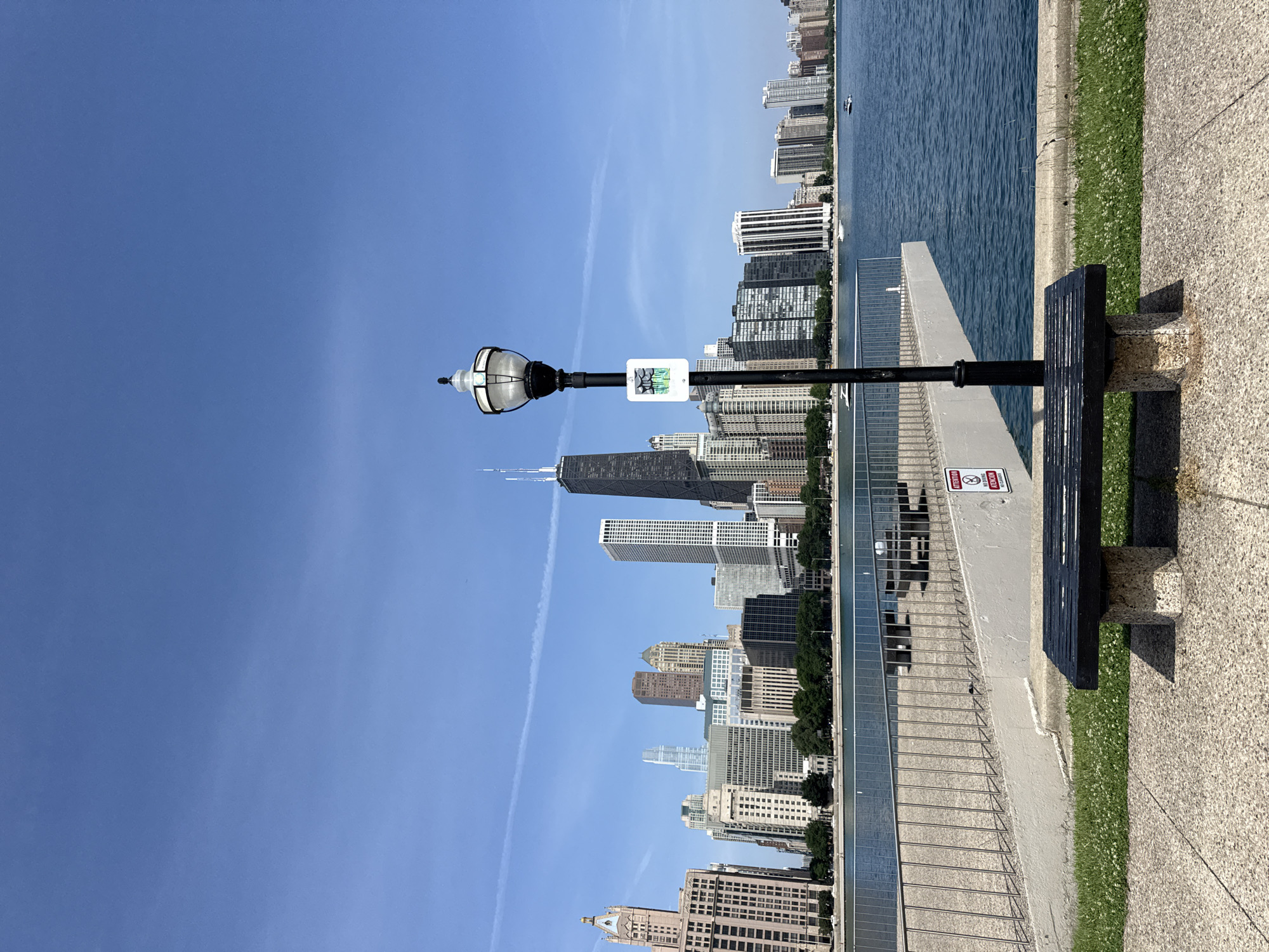 This media file captures a bright, sunny day in Chicago, United States of America. The scene looks out over the expansive blue waters of Lake Michigan, with a prominent skyline of diverse skyscrapers stretching across the horizon. In the foreground, a black park bench sits on a paved walkway, adjacent to a strip of green grass dotted with small white flowers. A lamppost stands beside the bench, supporting a light fixture and a small sign with a graphic image that appears to be a nature scene. The clear blue sky suggests it is midday. The image is oriented vertically, creating a dramatic perspective of the city and the lake. There is no visible activity or event taking place, presenting a peaceful, scenic view of the urban landscape meeting the natural environment. The overall mood is serene and picturesque.