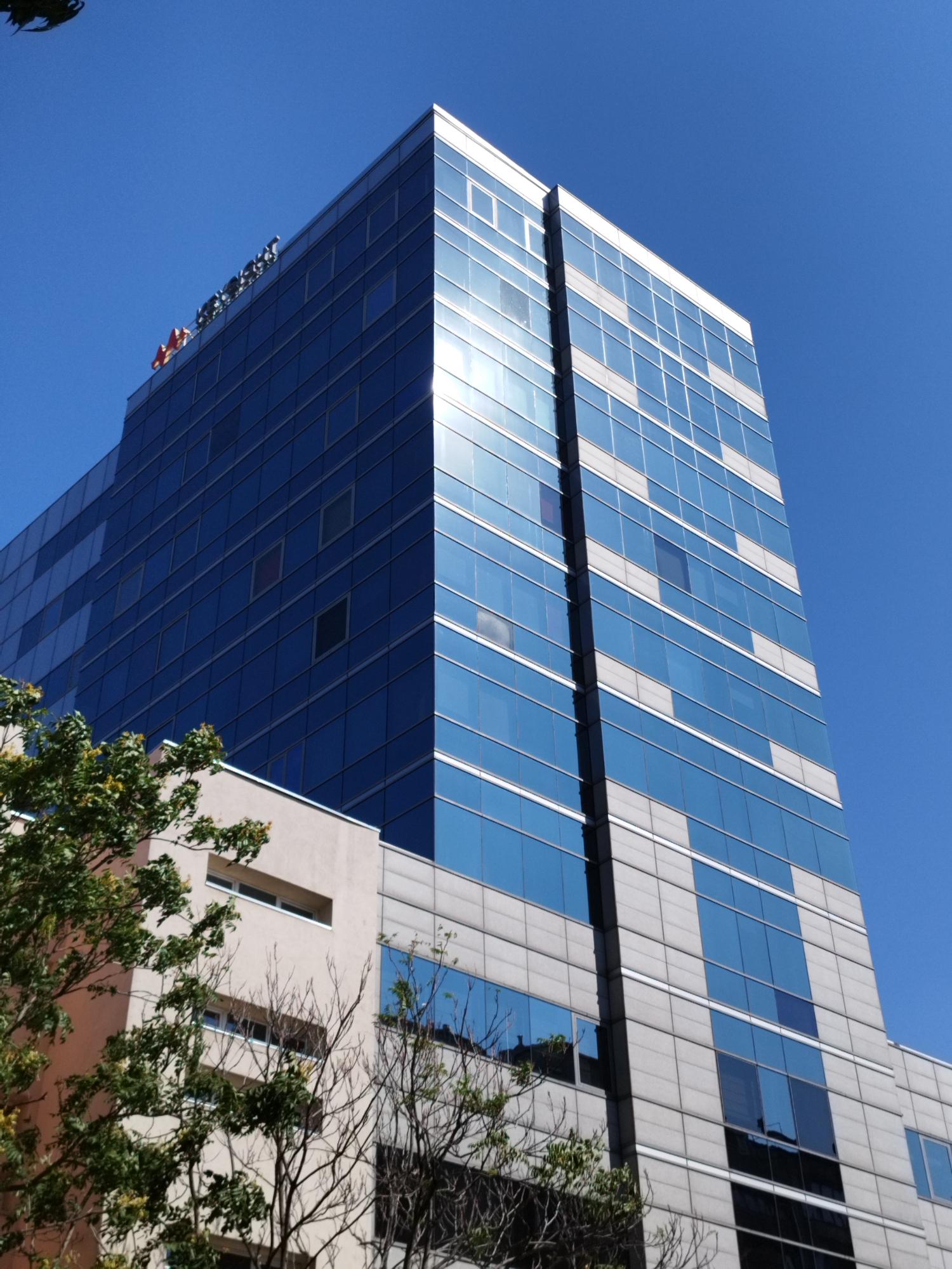 A tall, modern office building with a glass facade stands against a clear blue sky in Bucharest, Romania. The building features a combination of dark blue reflective glass panels and lighter gray concrete sections. A sign with red and white lettering is visible at the top of the building. Trees with green leaves are positioned in the foreground, partially obscuring the lower levels of a separate, lighter-colored building adjacent to the glass structure. The image is taken from a low angle, looking upwards.