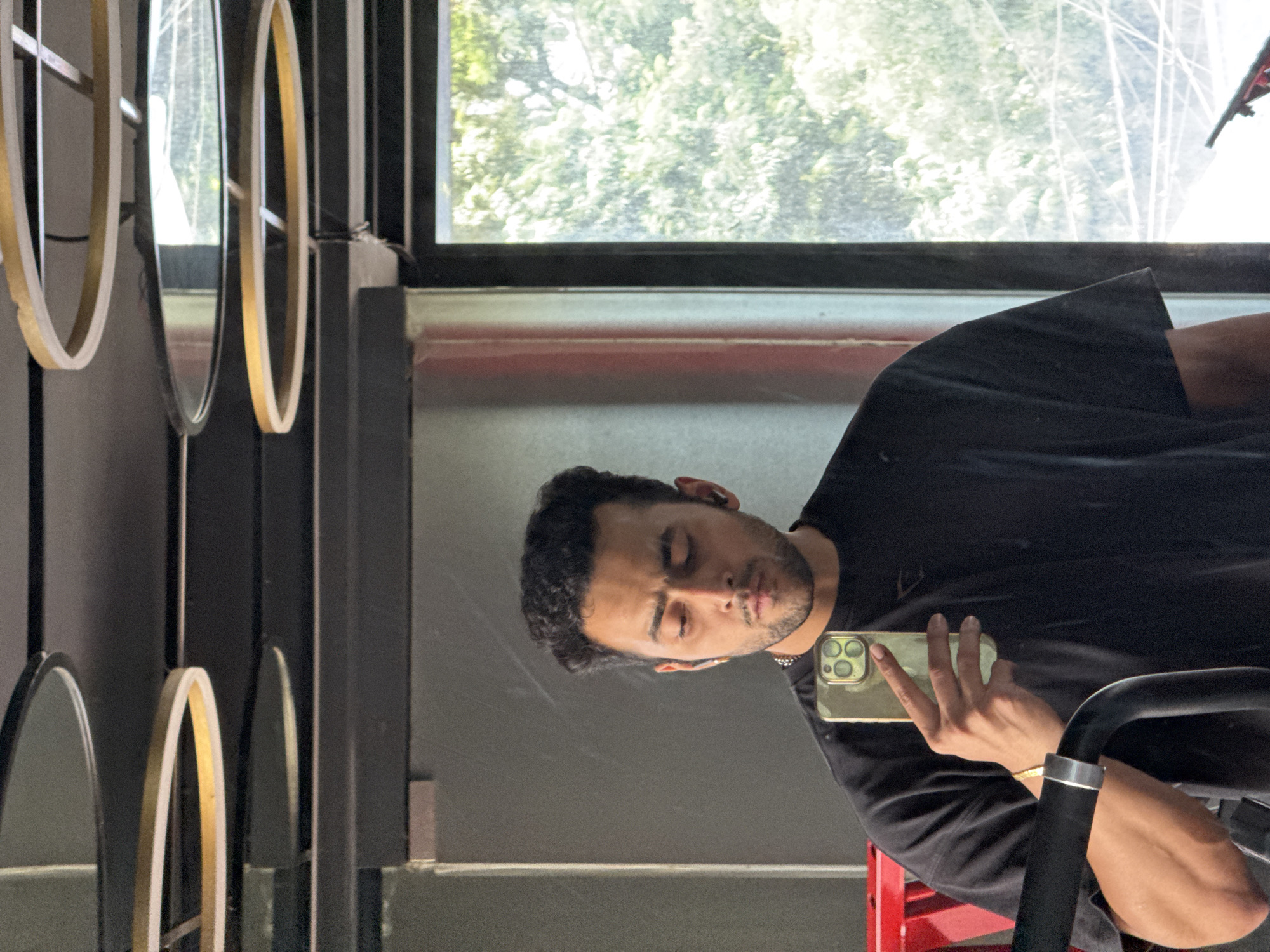 A man is taking a mirror selfie in a gym. He is wearing a black t-shirt and has short, dark, curly hair. He is looking down at his phone, which he is holding in his right hand. He has a slight beard and is wearing earbuds. In the background, there is a window with trees visible outside. The gym also has a ceiling with circular light fixtures. The location is not specified as Kolkata, India, in the provided image or crops.