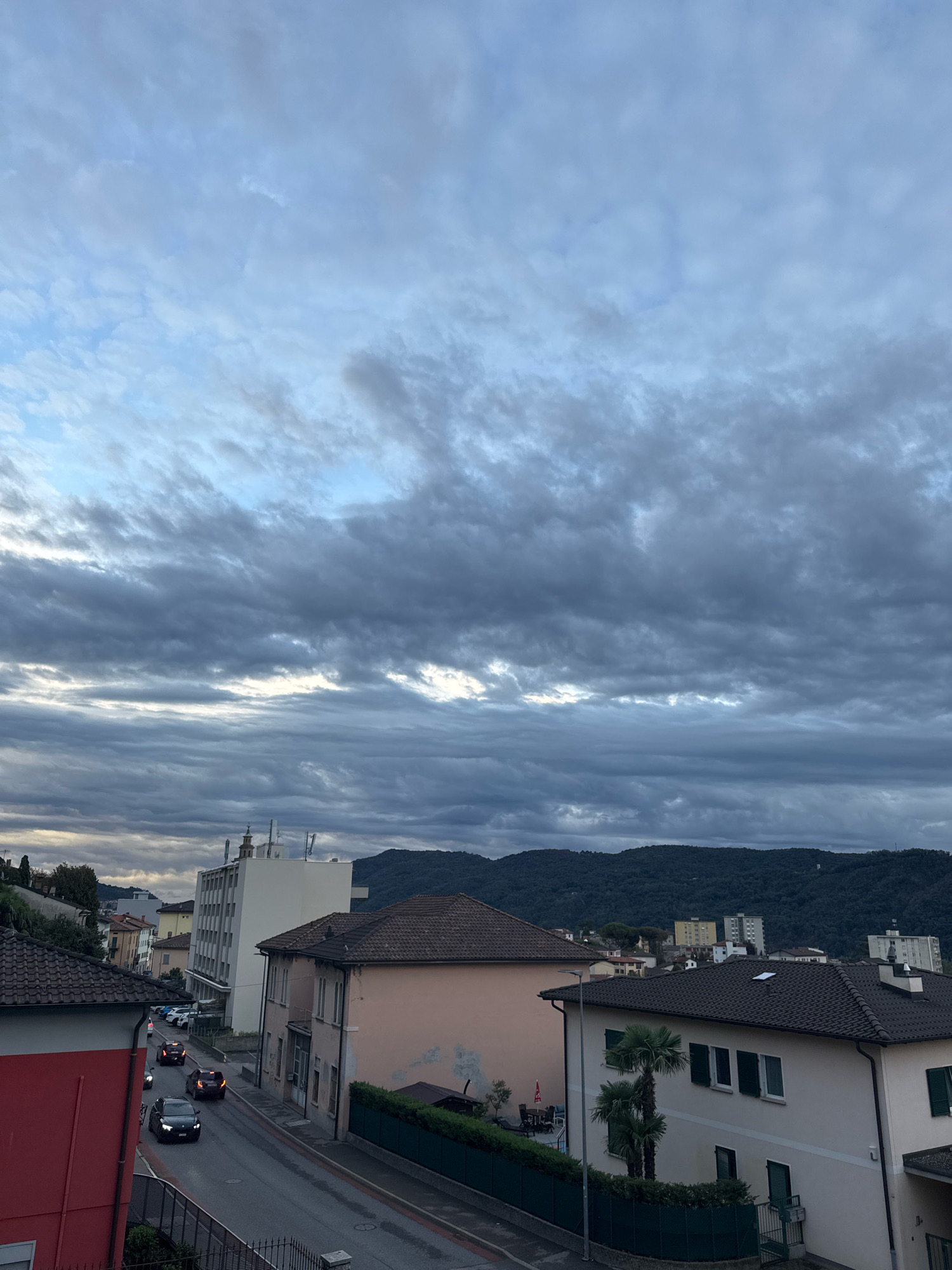 The image depicts an overcast sky over a residential area in Balerna, Switzerland. Dark, layered clouds dominate the upper portion of the frame. Below, a street curves through a neighborhood with various buildings. On the left, a red-sided building is partially visible. Further down the street, a light-colored building with multiple floors stands prominently. The street itself is wet, suggesting recent rain, and several cars are driving on it. To the right of the street, a pale pink building with dark tile roofing is visible, and beyond that, a light-colored building with green shutters and palm trees in front. In the background, rolling, tree-covered hills form the horizon.