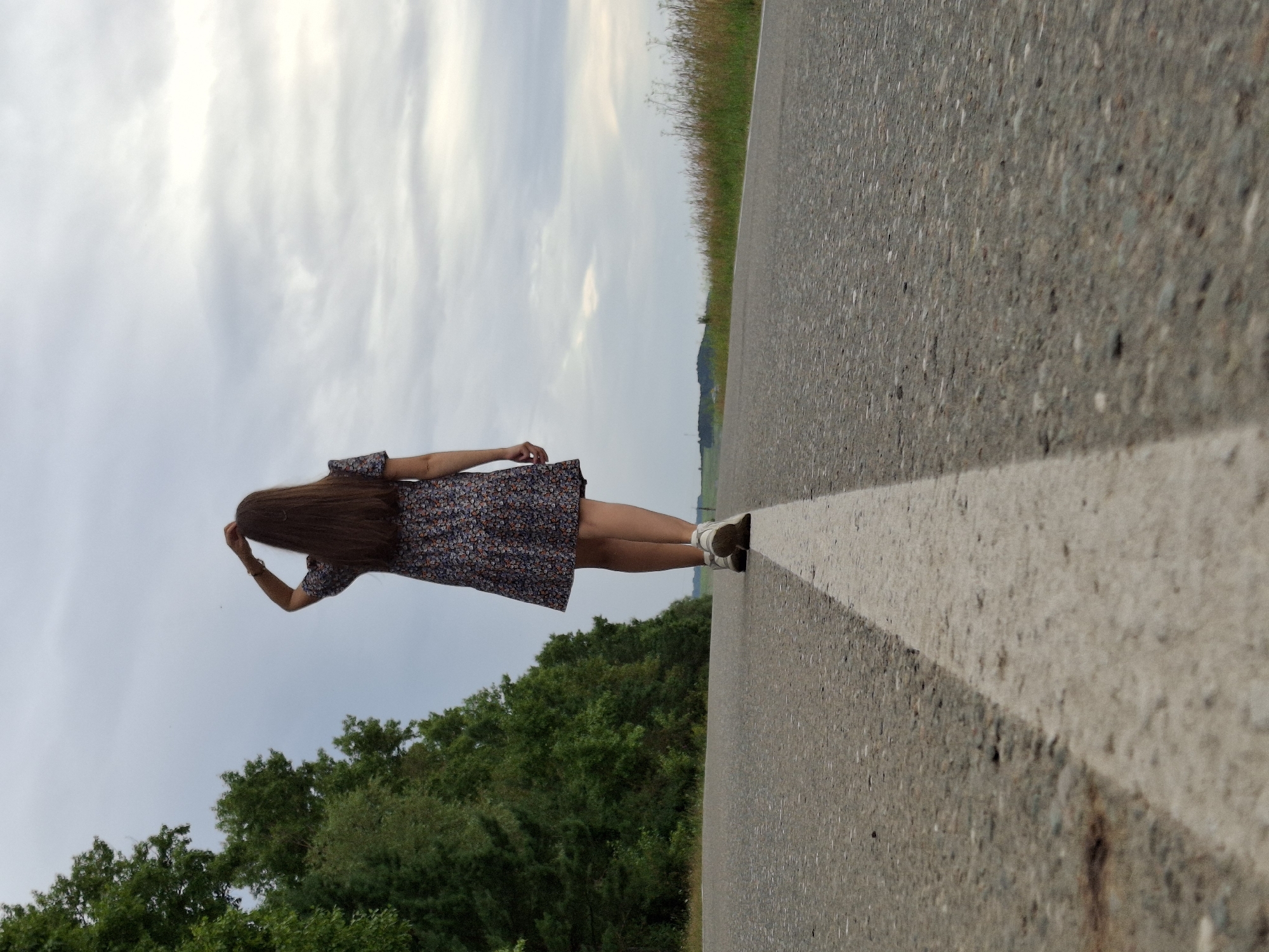 A person with long brown hair, wearing a floral print dress and white sneakers, is walking away from the camera on an asphalt road. The person is positioned in the center of the frame, with their right hand touching their head. A white line runs down the middle of the road, separating the lanes. To the left of the road is a dense line of green trees, and to the right, the road continues into a grassy field with some dried plants. The sky is overcast with gray clouds. The setting appears to be a rural area, possibly on the outskirts of the city of Garevaya, Russia, given the vegetation and the road conditions.
