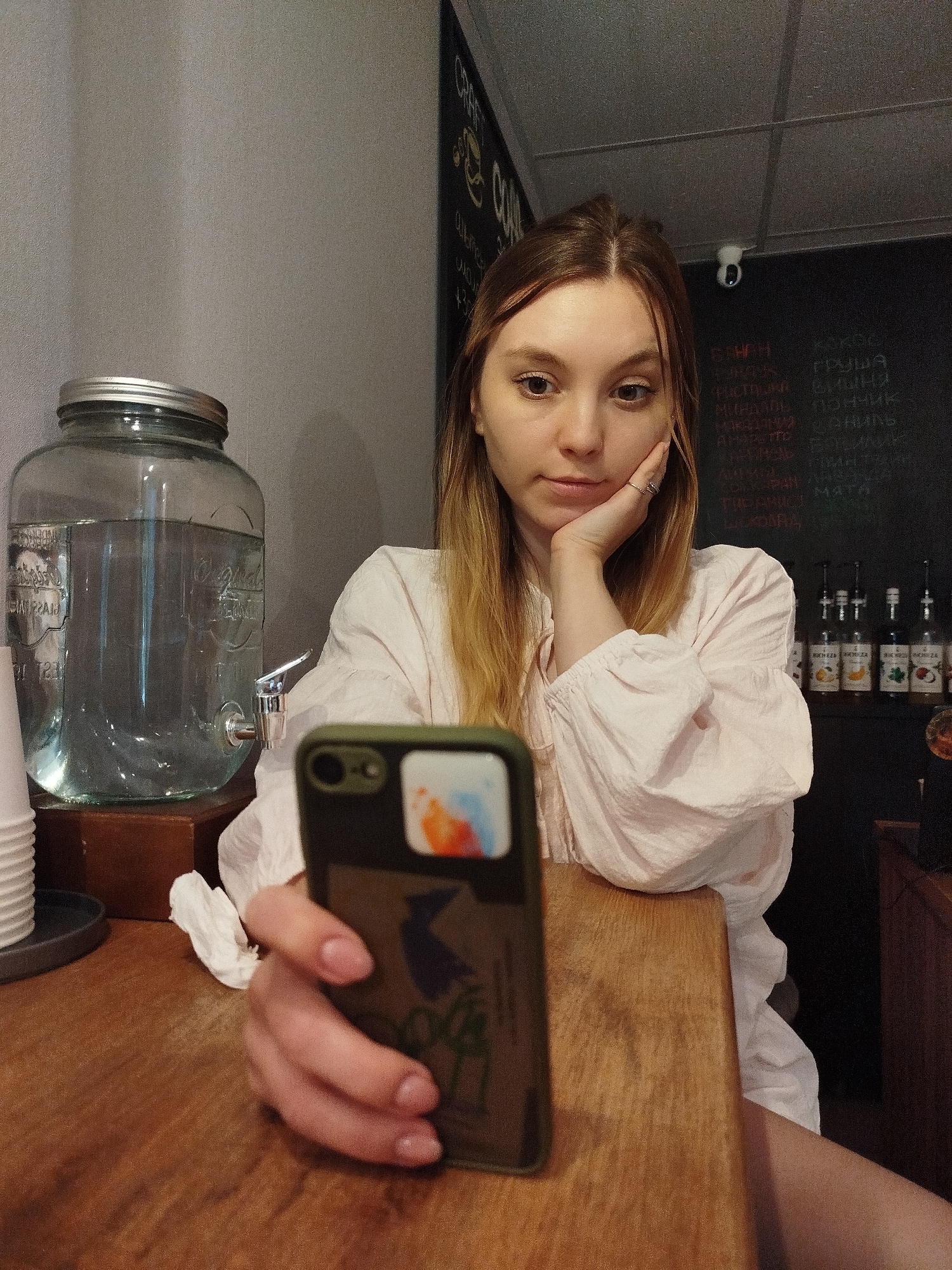 A young woman with light brown hair and fair skin is holding a smartphone in the foreground, looking at it with a neutral expression. She is resting her chin on her hand, which is adorned with a ring on her index finger. She is wearing a light pink, voluminous blouse with puffed sleeves. The background features a large glass jar filled with clear liquid, possibly water or a beverage, with a metal spigot at the bottom. To the right, a blackboard displays writing in white chalk, and a row of small bottles with labels is visible. The setting appears to be indoors, likely a cafe or a similar establishment, suggested by the blackboard menu and the bottles of syrups or flavorings. The lighting is dim, indicating it could be evening or a dimly lit indoor space. The woman's posture and gaze suggest she is engaged with her phone, possibly taking a selfie or browsing content.