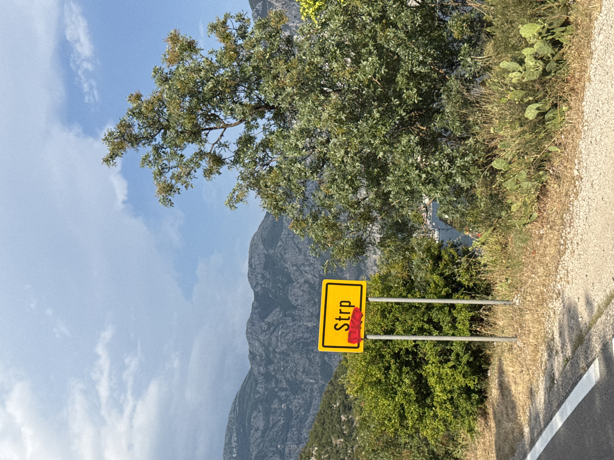 A yellow road sign with black lettering that reads "Strp" is positioned on the side of a road. The word "Strp" is partially obscured by a red painted rectangle. The sign is supported by two silver poles. The scene is set against a backdrop of a rugged mountain range under a partly cloudy sky. Lush green trees and dry brush surround the sign, and a body of water with boats is visible in the distance. The road in the foreground is paved and has a white line marking its edge. The location appears to be Strp, Montenegro, based on the sign.