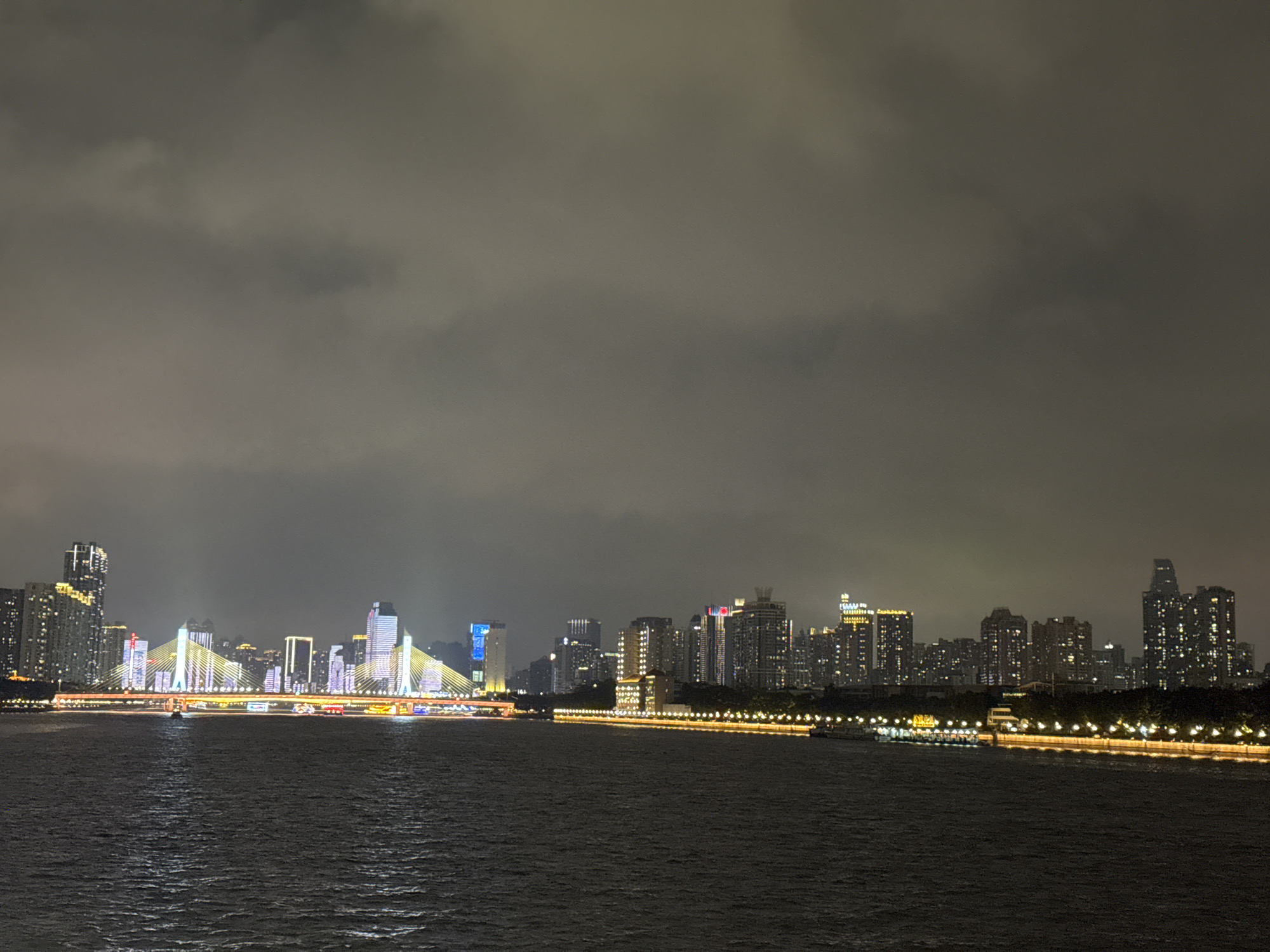 The image depicts a nighttime cityscape of Haizhu District, China, viewed across a body of water. The sky is overcast, and the city's skyline is punctuated by illuminated high-rise buildings and a cable-stayed bridge adorned with lights. Along the waterfront, a line of lights suggests a promenade. The water surface is dark with subtle ripples. There is a small boat in the water to the left of the image.