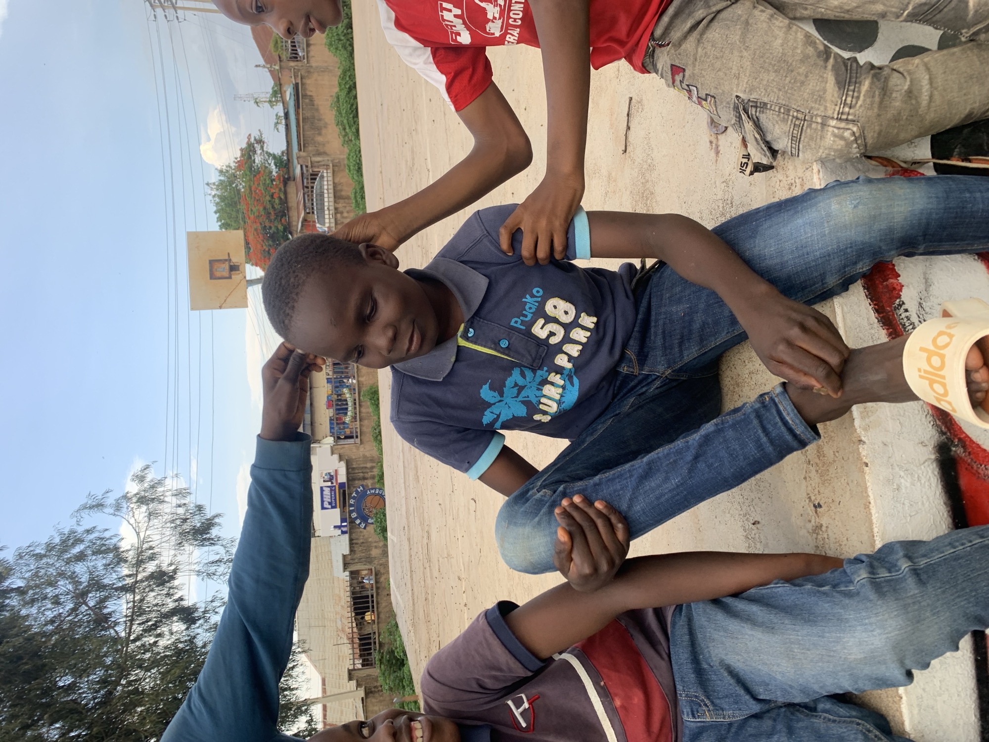 Four Black children are on a concrete surface outdoors. One child is seated, wearing a blue t-shirt and jeans. A child wearing a red t-shirt is partially visible. Another child with a blue long-sleeved shirt is visible only from the elbow up. The fourth child, wearing a maroon shirt, is partially visible on the bottom of the image. The seated child is the main subject. In the background, there is a basketball hoop, a building with business signs, trees, and a cloudy sky. The city is Jos, Nigeria.