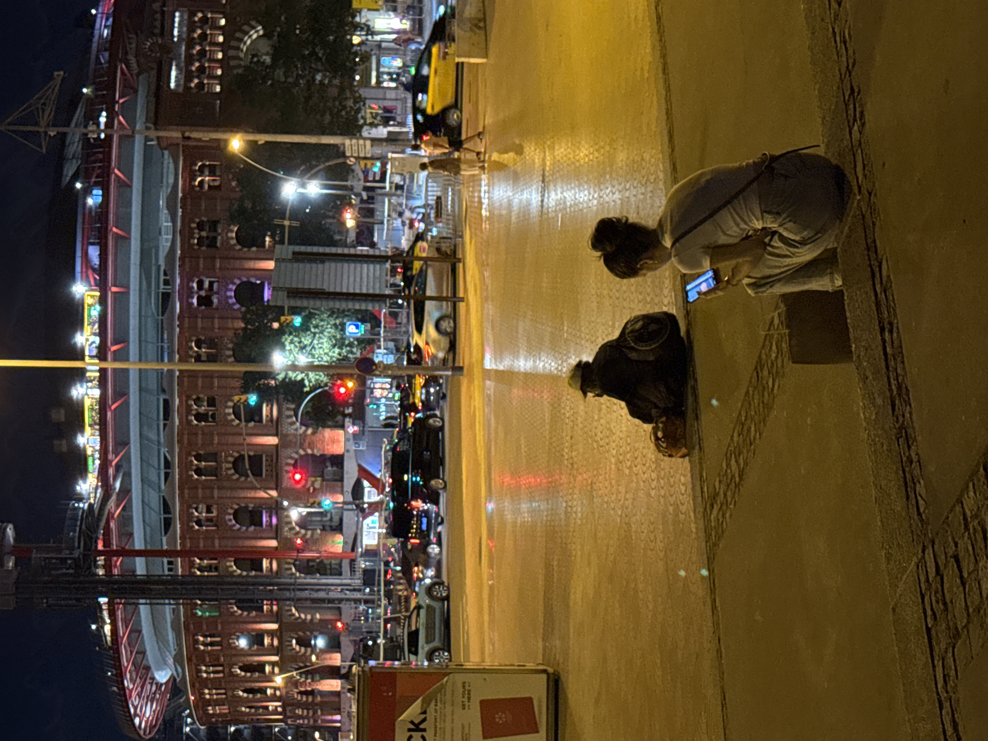 The image captures a nighttime street scene in Barcelona, Spain, with a large, illuminated building resembling a stadium or arena in the background. The foreground features a wet, reflective pavement, indicating recent rain or street cleaning. Two individuals are present in the foreground. On the left, a person wearing a dark hooded jacket and a hat is seated on the ground, facing away from the camera, with a backpack beside them. On the right, a young woman with her hair in a bun is sitting on a concrete ledge, looking at her smartphone. Several vehicles, including cars, SUVs, and a yellow taxi, are visible on the street in the mid-ground. Traffic lights and streetlights illuminate the scene. A promotional stand with the word "CK" is on the far left.