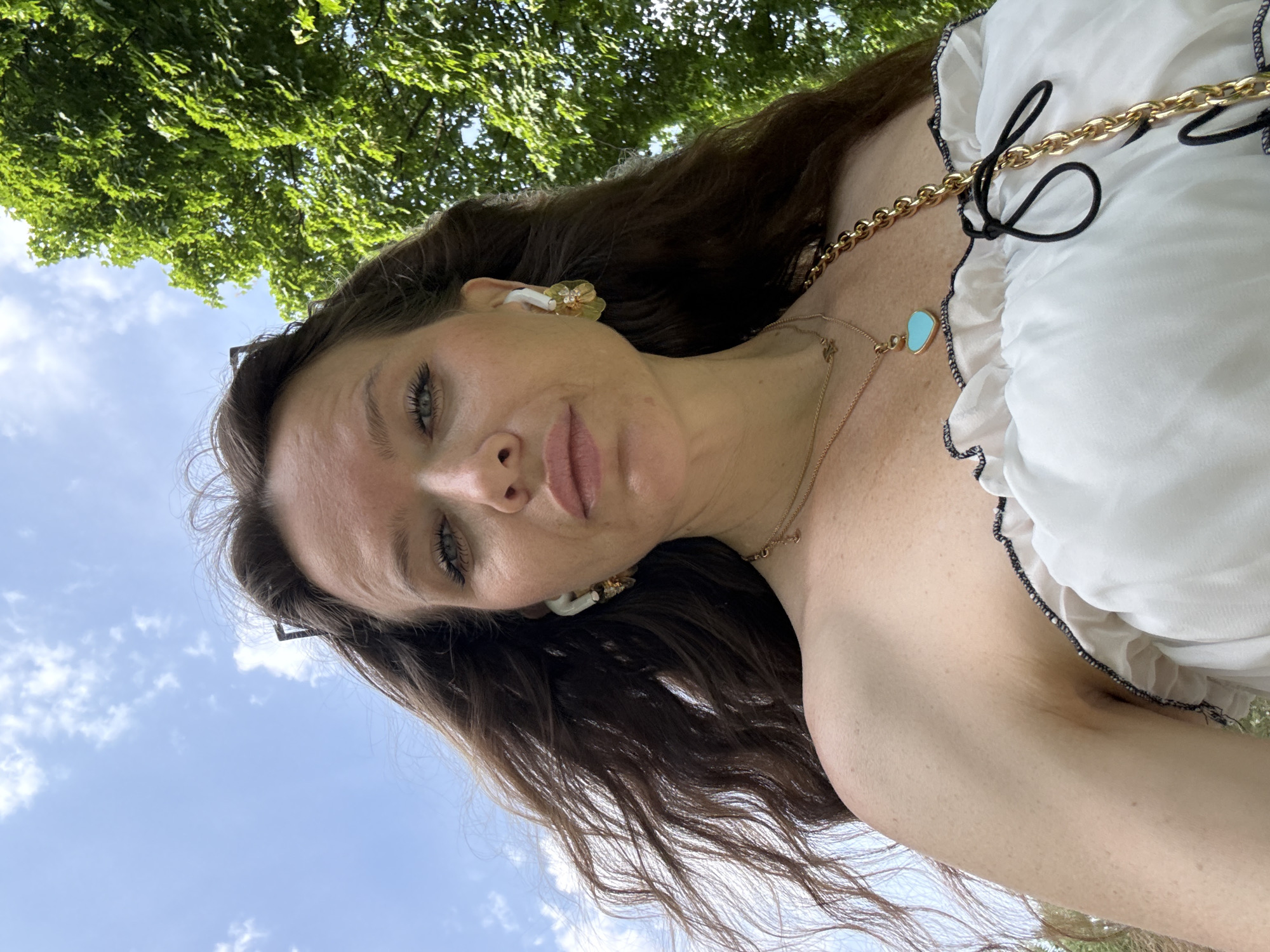 A woman is seen from a low angle against a backdrop of a tree and sky. She has brown hair, fair skin, and is wearing a white top with black trim. She has a gold chain necklace and earrings. The location is unclear, and there is no visible evidence suggesting that this image was taken in Moscow, Russia.
