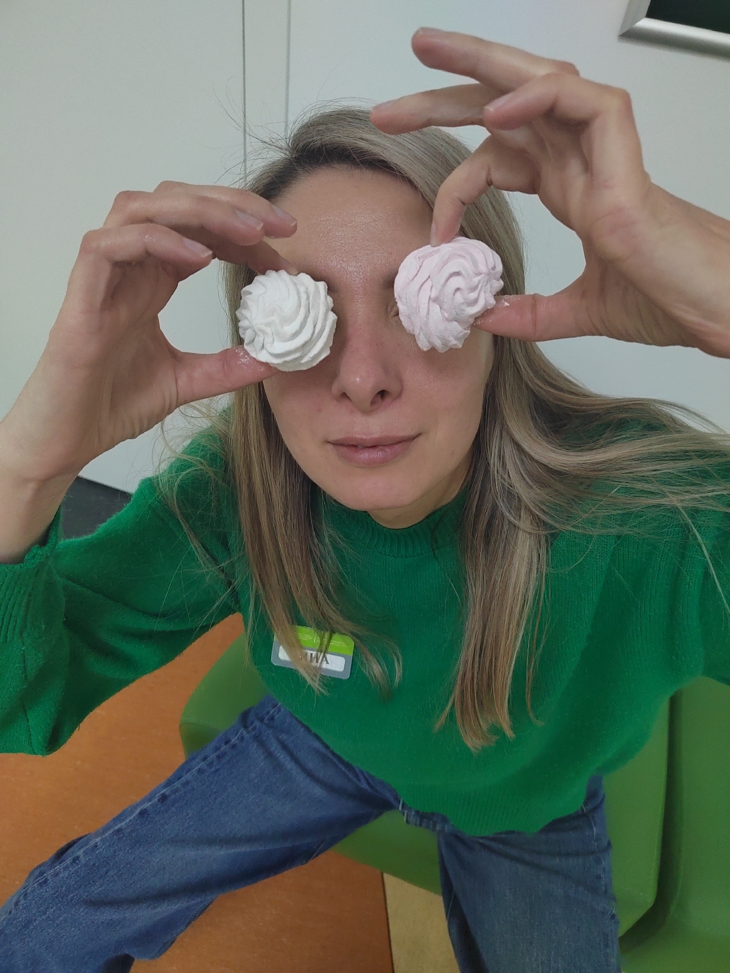 A fair-skinned woman with blonde hair is seated indoors, likely on a green chair situated on an orange floor. She is wearing a green sweater and blue jeans. She is holding a white meringue in her right hand and a pink meringue in her left, positioning them over her eyes. The environment includes a white wall. Based on the location metadata (Votkinsk, Russia), this scene presumably occurs somewhere in that city.