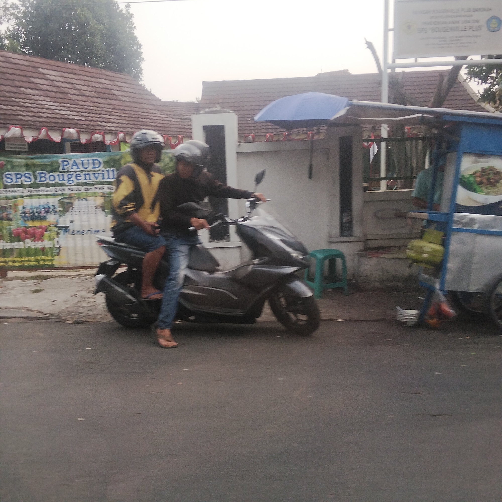 In Bogor, Indonesia, two individuals are riding a dark-colored scooter on a paved road. The person in the front is wearing a dark-colored helmet and a black jacket, while the person in the back is wearing a yellow and black jacket and a gray helmet. Both are wearing casual clothing.
Behind them, a banner with text that appears to be in Indonesian is visible. The banner has red and white decorative elements along its top edge. Further in the background, a building with a red-tiled roof is present, along with trees and a blue umbrella. A food cart with a blue frame and a silver body, containing a gas cylinder, is parked on the right side of the road. A white bowl with a lid is on the ground near the food cart. The sky is overcast.