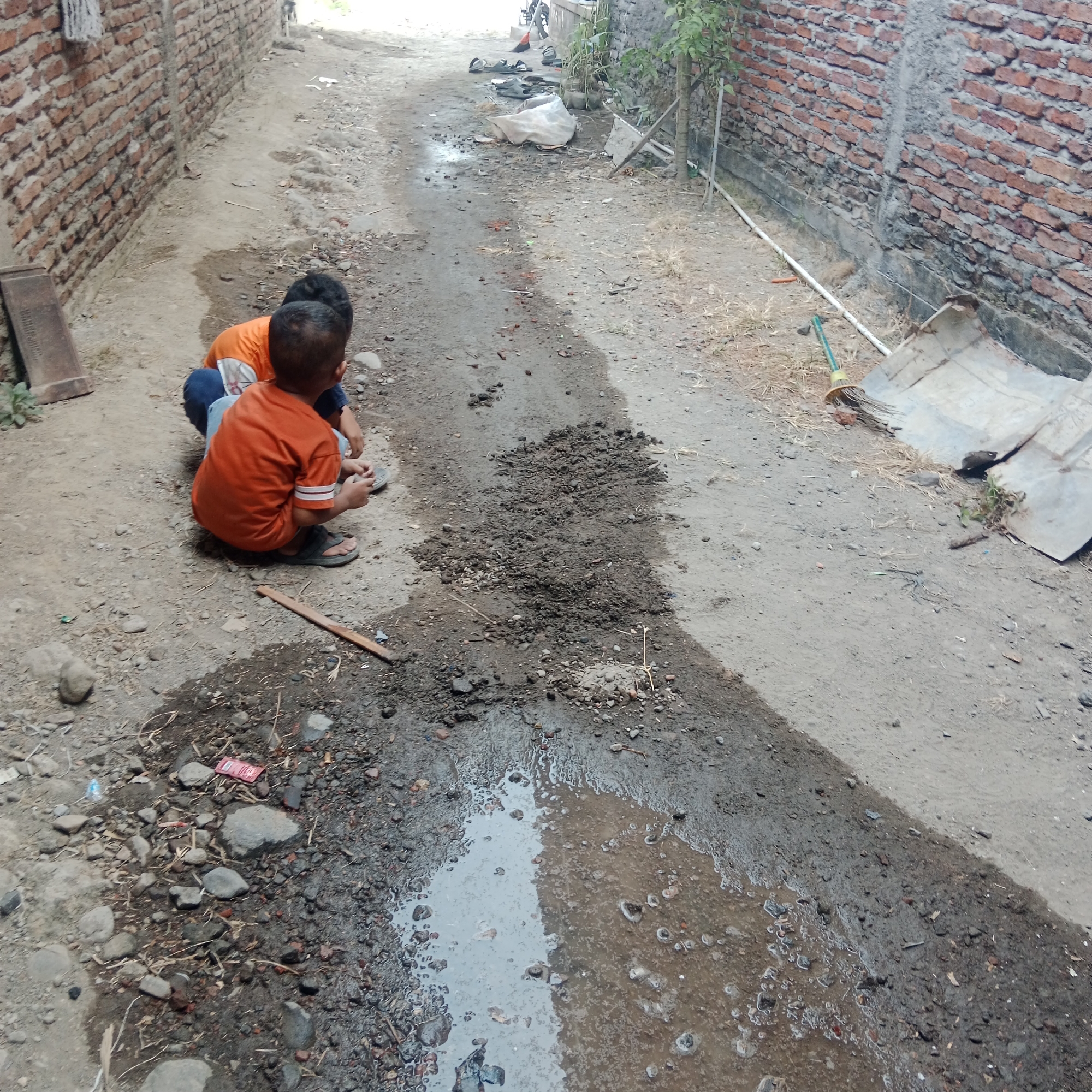 The image depicts an outdoor scene, likely an alleyway or path, in Tegowanu Kulon, Indonesia. Two young children, dressed in casual clothing, are interacting with the ground. One child, wearing an orange shirt and dark pants, is kneeling and appears to be playing with mud or dirt. Another child, also in an orange shirt, is positioned behind them, their head visible. The ground is a mixture of dirt, gravel, and patches of water, suggesting recent rain or a leaky water source. The environment includes a brick wall on the left side of the image, a similar wall on the right, and vegetation in the background. Scattered objects like rocks, debris, and a wooden stick are also present on the ground. The overall impression is one of children engaging in simple outdoor play in a somewhat unkempt urban or semi-urban setting.
