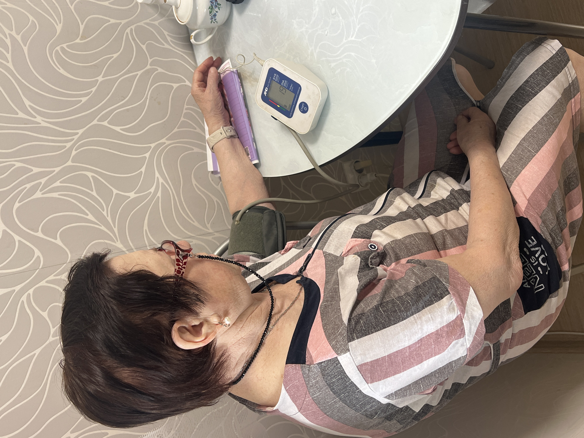 An elderly woman with short, dark hair is seated indoors at a light-colored table, engaged in taking her blood pressure. She is seen in profile, wearing eyeglasses with a beaded chain around her neck and a casual, striped top featuring pink, white, and grey/brown hues. A watch is visible on her left wrist.
Her left arm is fitted with a blood pressure cuff, connected to a white and blue A&D brand automatic blood pressure monitor resting on the table. The monitor's screen displays numerical readings, including "119" for SYS (systolic) and "69" for DIA (diastolic), and "73" for "ПУЛЬС" (PULSE), with "СТАРТ" (START) button visible. In her right hand, she holds a purple-covered notebook or pamphlet.
On the table near her, white porcelain teacups or teapots with delicate blue floral patterns are stacked. The wall behind her features light grey wallpaper with a flowing, organic white pattern. Part of her striped clothing also shows faint text, where "IS LOVE" is discernible. The setting appears to be a home environment in Perm, Russia, given the context provided. The woman's expression is neutral, suggesting focus on the activity.