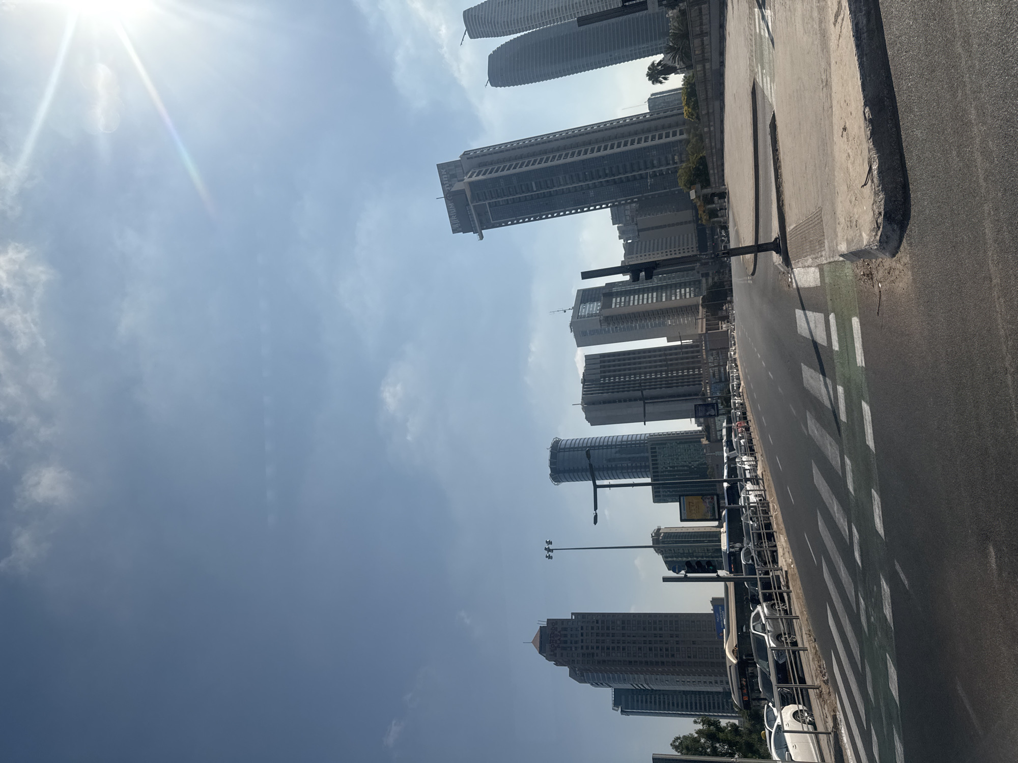 This is a ground-level, wide-angle photograph taken on a sunny day in Tel Aviv, Israel, facing north. The foreground features a paved road with a prominent pedestrian crossing painted in white and green stripes. To the left of the road, a line of parked cars, including white sedans and darker SUVs, are visible behind a metal railing. Beyond the parked cars, several buses are also lined up. In the background, a dense cluster of modern high-rise buildings dominates the skyline. The sky is a clear blue with a few wispy clouds and a bright sunbeam radiating from the upper right corner. The buildings vary in height and architectural style, with some appearing sleek and glass-fronted, while others are more solid and rectangular. One of the taller buildings has "LEONARDO PLAZA" visible on its top. Another prominent building, tilted slightly in the frame, displays "ATRIUM MINK" signage. The scene depicts a typical urban environment in Tel Aviv, characterized by its contemporary architecture and busy street life.