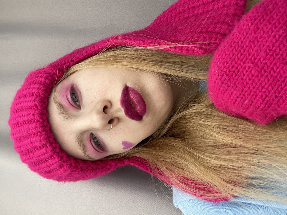 A young adult female is depicted in a head-and-shoulders portrait, leaning against a plain light grey wall in Izhevsk, Russia. She has fair skin, long blonde hair, and is looking directly at the viewer. Her eyes are accentuated with bright pink eyeshadow, extending to the lower lash line. Her lips are coated with dark pink/fuchsia matte lipstick. A small, light purple heart decal or paint is present on her left cheek, below the eye. A small silver-colored hoop piercing is visible in her left nostril. She wears a thick, ribbed, bright fuchsia pink knitted hat, which appears to have extended ear flaps or similar features, and a matching knitted mitten is visible in the lower right of the frame. A light blue, possibly knitted or textured, long-sleeved top is partially visible on her left shoulder and arm.