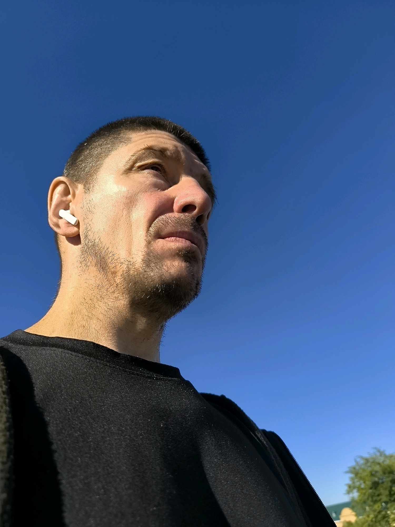 The image shows a man from the chest up, looking upwards against a clear blue sky. He has short, dark hair and stubble on his chin and jawline. He is wearing a black t-shirt and white wireless earbuds in his left ear. The perspective is from a low angle, emphasizing the sky. The background shows a blurry green tree on the right side and some indistinct structures. There is no visible indication of the Qarağandı city administration, Kazakhstan.