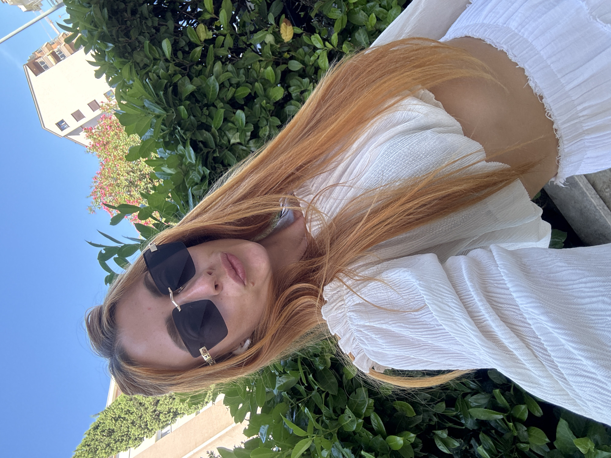 A red-haired woman wearing sunglasses and a white crop top stands outdoors, with a lush green hedge and a building in the background. The woman is angled towards the camera, with her head slightly tilted and her lips pursed. She has long, wavy red hair that falls around her shoulders and a white ruffled top that exposes her midriff. The sunglasses are black with a gold frame. The hedge is dense and green, with some pink flowers visible behind it. The building appears to be a multi-story structure, likely residential, with balconies. The sky is a clear blue. The scene suggests a warm, sunny day, possibly in a Mediterranean setting like Budva, Montenegro, known for its coastal beauty and similar architecture.