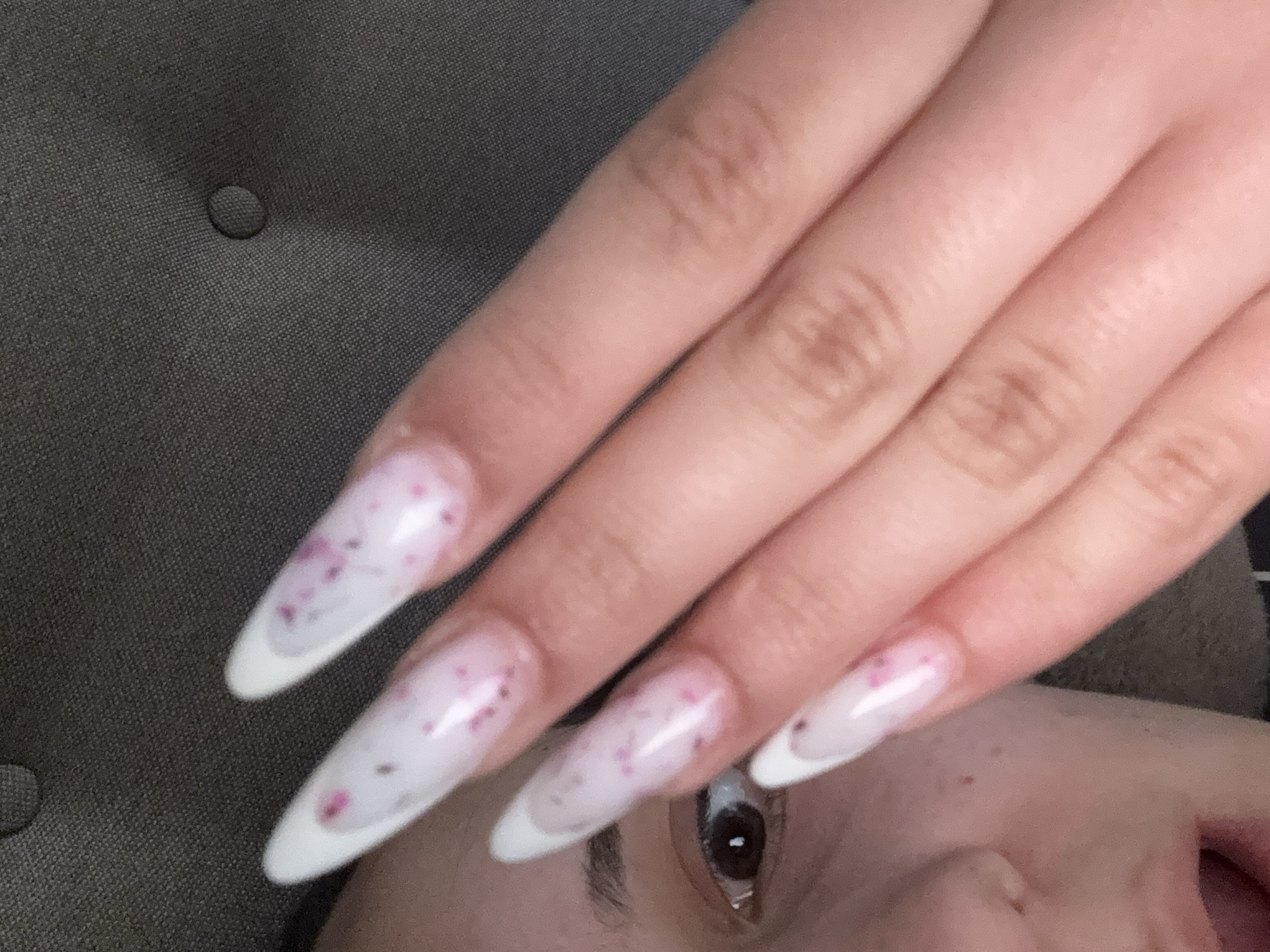The image is a close-up, vertical, slightly blurry shot of a person's left hand with long, pointed, acrylic nails adorned with pink speckles and a white tip, and a portion of their face. The nails are applied over a light pink base. The person's left eye, eyebrow, and part of their mouth are visible on the left side of the frame. The background appears to be a textured, dark gray fabric, possibly upholstery. The lighting is soft and diffused, creating subtle shadows. The scene is indoors, with no discernible city or location details.