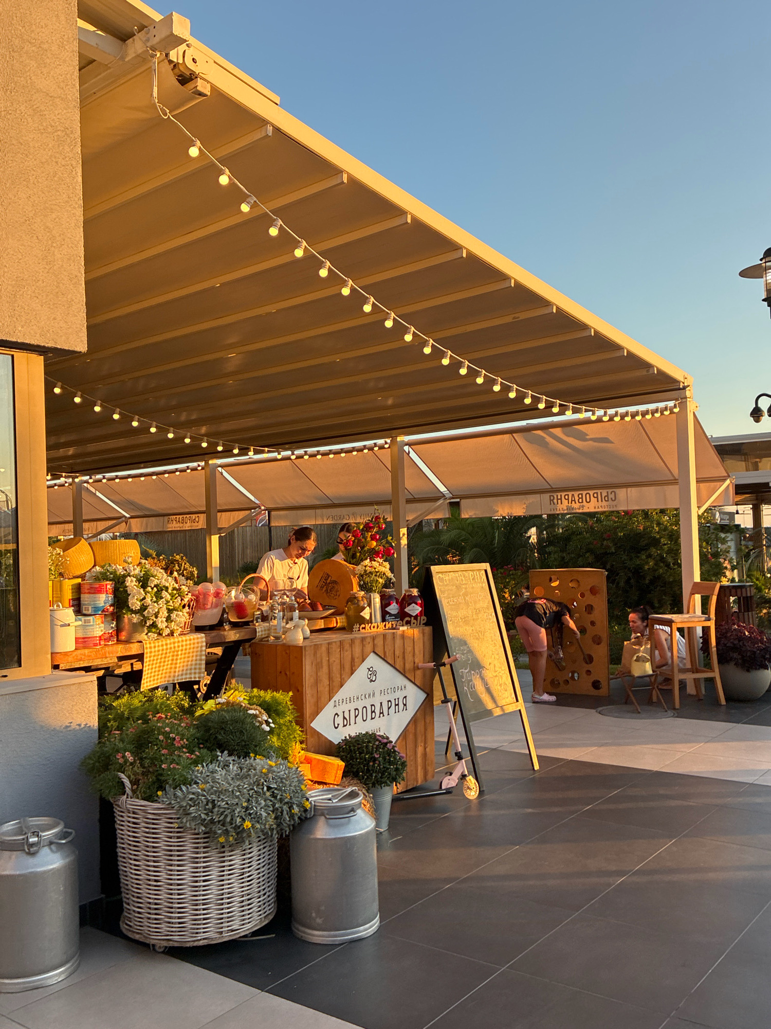 The image depicts an outdoor market stall in Sochi, Russia, during sunset. A string of illuminated bulbs hangs from a retractable awning, casting a warm glow over the scene. The stall is laden with various items, including baskets of flowers, jars of preserves, and large wheels of cheese. Two individuals are visible: a vendor preparing food at the stall and a customer interacting with a large, cheese-themed game. In the background, lush greenery and other market structures are visible, suggesting a lively outdoor gathering. The ground is paved with dark tiles, and the overall atmosphere is relaxed and inviting, characteristic of a pleasant evening at a local market.