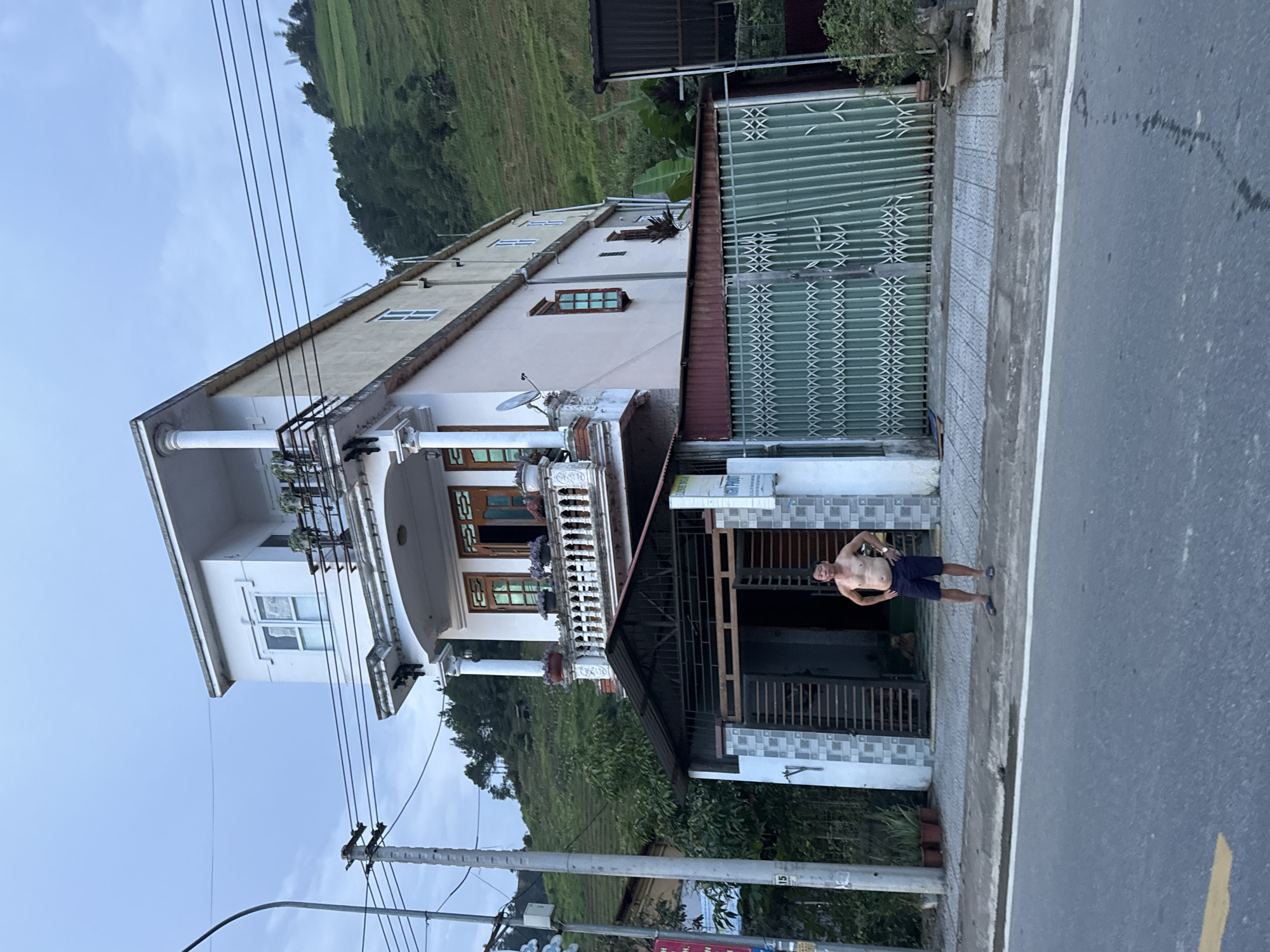 The image, when viewed upright, displays a street scene in a rural or semi-urban setting, consistent with the Mù Cang Chải region of Vietnam.
The primary subject is a multi-story house, painted in light shades of white and beige, featuring multiple balconies with ornate white balustrades and columns. A satellite dish is mounted on an upper balcony. The ground floor entrance is protected by dark metal grates, and next to it, a large green metal gate with a diamond-patterned lattice is visible.
On the sidewalk in front of the house, a shirtless man stands facing the camera, hands on his hips. He is wearing dark shorts and blue flip-flops, appearing relaxed. The sidewalk is paved with patterned concrete tiles, and a gray asphalt road runs alongside it in the foreground.
In the background, behind the house, extensive green hills covered in agricultural terraces, likely rice paddies, stretch across the landscape, confirming the location context. Overhead, multiple power lines crisscross the sky, supported by a utility pole marked with the number "15".
The lighting suggests it is daytime, possibly overcast or late afternoon, with a light blue to gray sky. A small sign near the house's ground floor entrance is partially visible, showing text that includes "HOMESTAY GUEST HOUSE" and "SƯƠNG MÙ," indicating it is likely a local accommodation named Sương Mù Homestay.