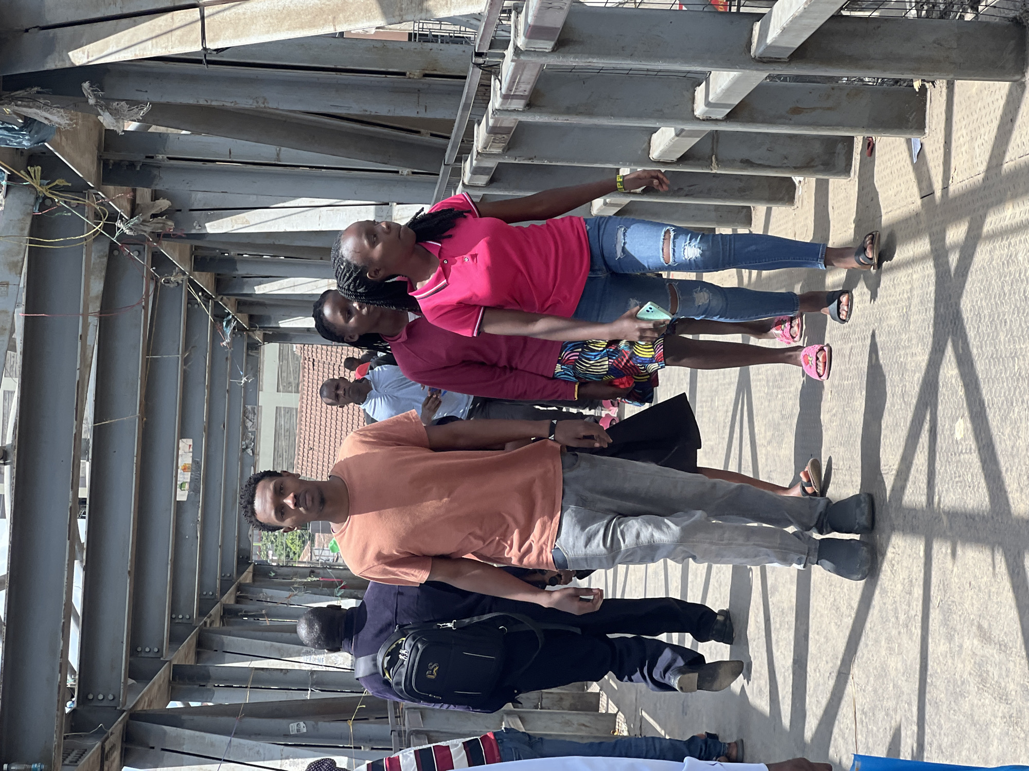 The image depicts a group of individuals standing on a pedestrian walkway constructed of steel beams. The location appears to be in Nairobi, Kenya, based on the architectural style of the building visible in the background.
There are at least six individuals visible in the image. One person in the foreground is wearing an orange t-shirt and light gray pants. Another person is wearing a dark blue jacket and carrying a black backpack. Further back in the line, one person is wearing a pink polo shirt, another wears a red top, and a man is wearing a blue shirt.
The walkway is made of intersecting steel beams, casting shadows onto the concrete surface. There are wires and debris hanging from the beams above. A few buildings are visible in the background.