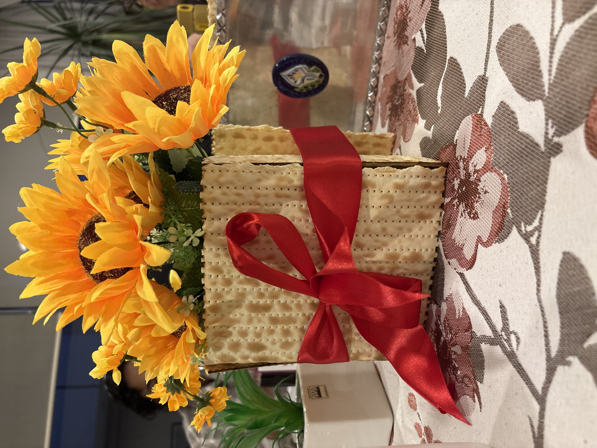 A stack of matzah is tied with a red ribbon and surrounded by yellow sunflowers and greenery. The matzah is on a tray or table with a floral patterned tablecloth. The location is Huixquilucan de Degollado, Mexico.
