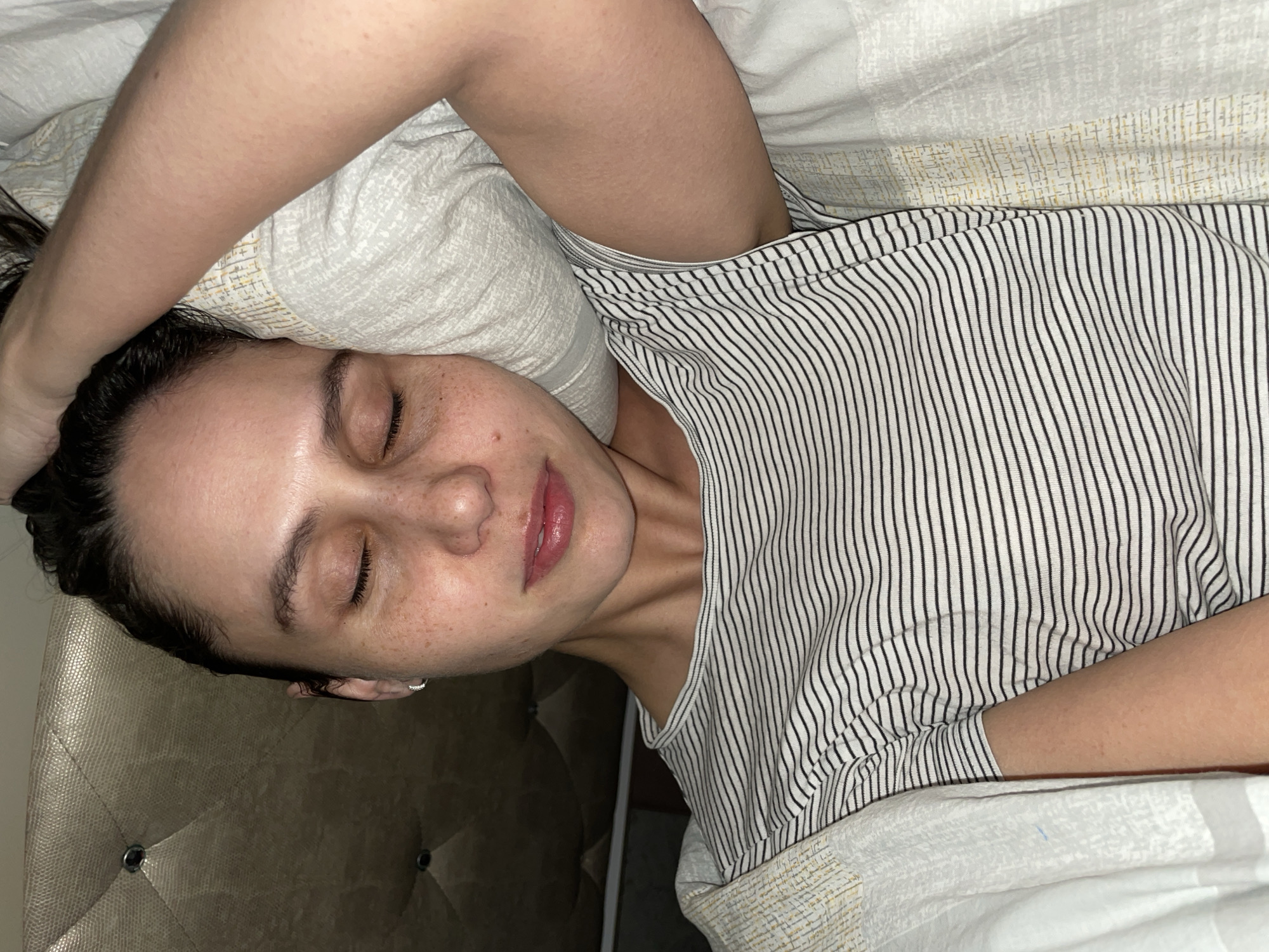 A young woman is lying in bed with her eyes closed. She is wearing a striped shirt. Her left arm is bent and resting on her forehead, which is touching the pillow. The bedsheets appear patterned. Part of a headboard can be seen. There is no indication this image was taken in Izhevsk, Russia.