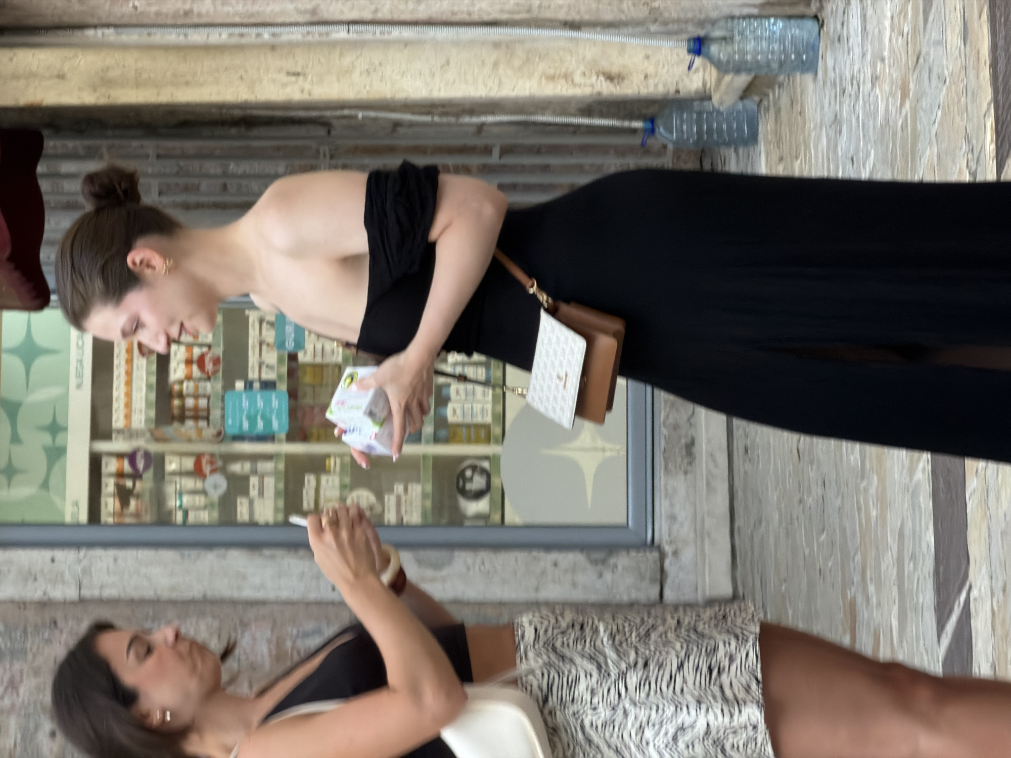 Two young women are standing outside a shop in Budva, Montenegro. The woman on the left, with short dark hair, wears a black crop top and a patterned skirt. She is holding a mobile phone and reaching out to the other woman. The woman on the right, with her hair in a bun, is wearing a black off-the-shoulder dress and holding a small white box. She has a small brown and white crossbody bag. The shop behind them appears to be a pharmacy with shelves of products visible through the glass. The street is paved with stone.