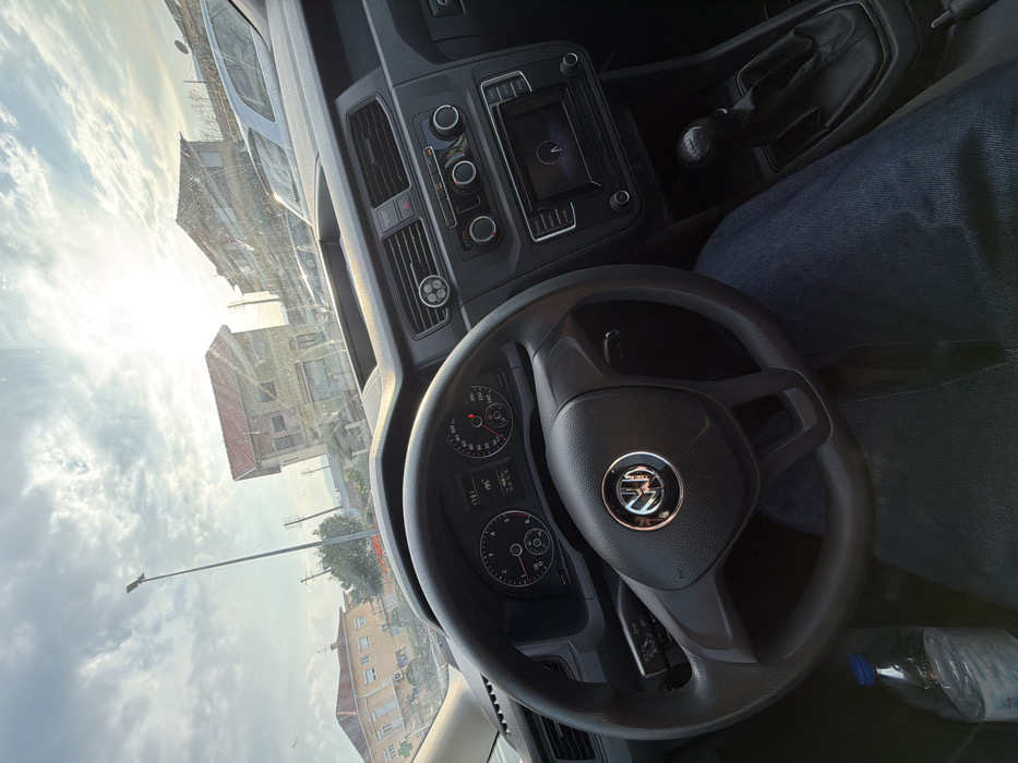 The image depicts an interior-exterior view from the driver's seat of a Volkswagen vehicle, likely a Caddy, located in Felgueiras, Portugal.
**Interior:** The foreground is dominated by the black three-spoke Volkswagen steering wheel. Behind it, the instrument cluster displays a digital time of 13:31 and an ambient temperature of 19.5°C. Analog gauges for engine RPM, speed, and fuel level are visible. The dashboard is dark grey, featuring central air vents, one of which has a small circular, three-pronged accessory clipped into it. Below the vents are controls for the climate system and audio, and a central infotainment screen that appears to be active or loading. To the right, a manual gear shifter is visible. The lower left shows a clear plastic water bottle with a blue cap resting on the floor or seat. The driver's legs, clad in blue denim jeans, are visible below the steering wheel and extending to the right.
**Exterior:** Through the windshield, the scene is illuminated by strong sunlight from the upper right, resulting in significant lens flare and glare. The sky is partly cloudy with patches of bright blue. In the mid-ground, a two-story building with a reddish-brown tiled roof and light-colored, possibly worn, facades stands prominent, slightly right of center. To its left, another multi-story building with a light beige or cream facade and a tiled roof is partially visible. A tall, dark street light pole is positioned to the left of the central building. In the background, parked vehicles and additional structures are discernible, indicating an urban or semi-urban environment. Utility poles and lines are also present in the distance. The road surface or parking area appears to be asphalt.