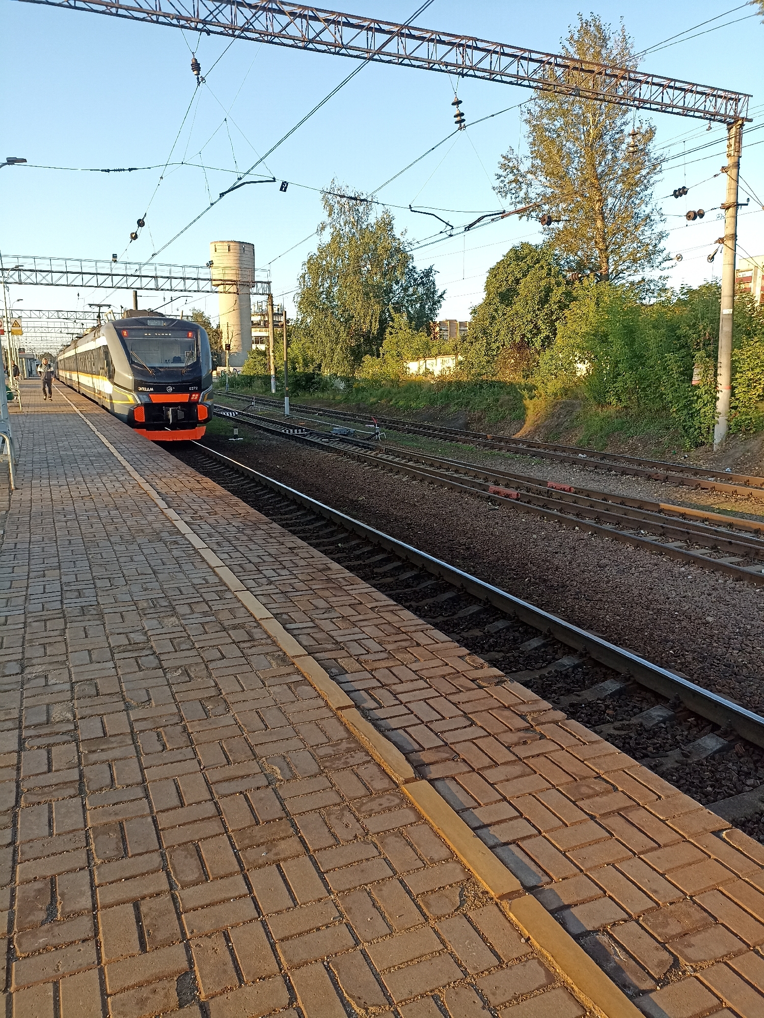 A modern, dark gray and orange electric train, numbered 6272, is stopped at a train station platform. The train has headlights on and its doors are closed. The platform is paved with light brown bricks and has a yellow safety line along its edge. To the left of the train, a person is walking away from the camera on the platform. In the background, there are multiple train tracks, overhead power lines supported by metal and concrete structures, dense green trees, and a tall cylindrical water tower. A few buildings are visible in the distance. The sky is clear and blue. The scene appears to be set during daytime, with the sun casting long shadows. The location is a train station in Chekhov, Russia.