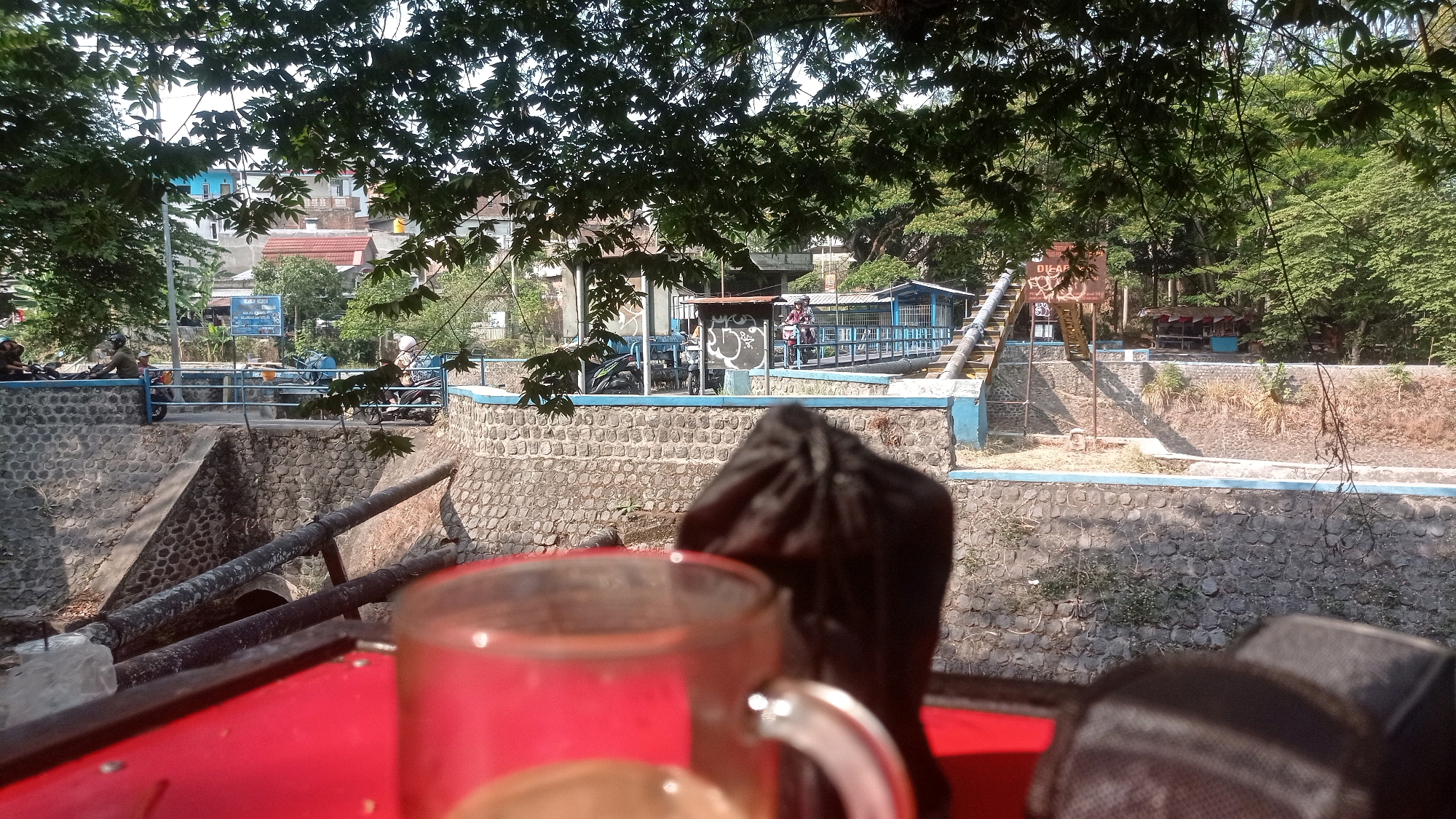 The image depicts a scene in Mulyoagung, Indonesia, likely near a water channel or canal. In the foreground, a red surface, possibly a table, is visible with a blurry glass mug and a dark fabric object. Beyond the foreground, a stone wall lines a channel. In the mid-ground, several large pipes run diagonally across the channel. A walkway or bridge, lined with a blue railing, spans the channel. On the walkway, individuals on motorcycles are visible, some appearing to be interacting. Buildings and trees are situated in the background, suggesting an urban or semi-urban environment. The overall atmosphere is one of daytime activity in an outdoor setting.
