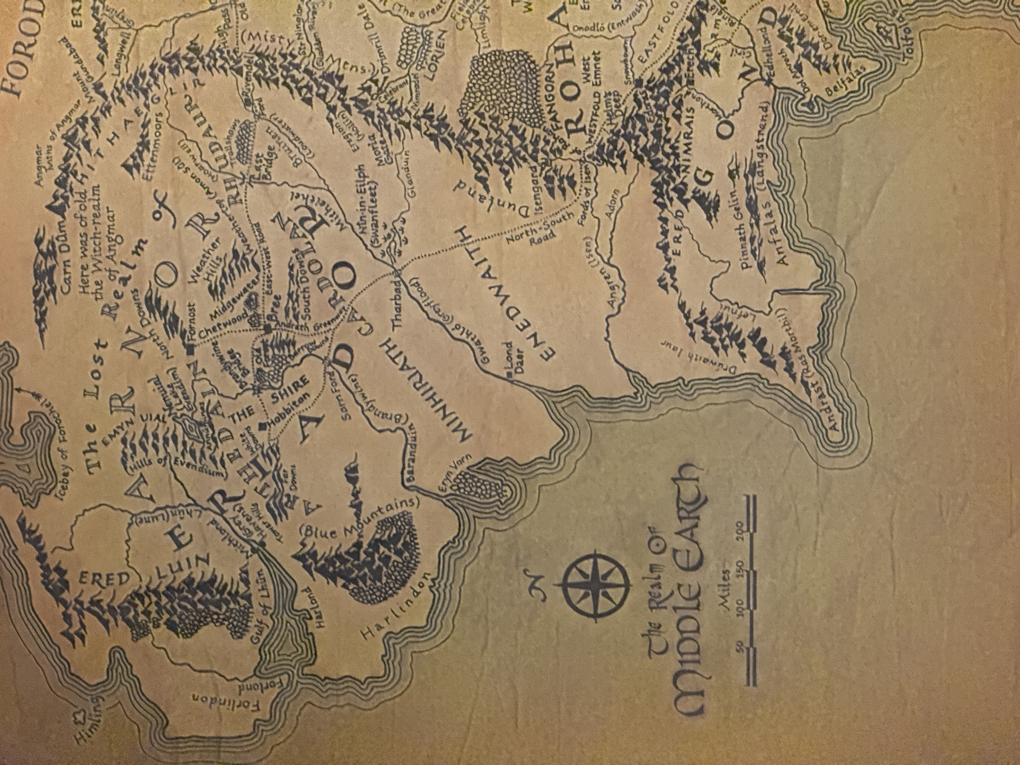 The image is a map of Middle Earth, the fictional setting of J.R.R. Tolkien's novels "The Hobbit" and "The Lord of the Rings". The map shows various regions, including the Shire, Eriador, Rhudaur, Cardolan, Minhiriath, Enedwaith, Rohan, Gondor, Ered Luin, and Forodwaith. Key landmarks such as the Misty Mountains, the River Anduin, and specific locations like Rivendell, the Brandywine Bridge, and the Fords of Isen are also marked. A compass rose indicating direction is visible in the lower center, and a scale for measuring distances in miles is provided. The style of the map evokes a hand-drawn, ancient document, with faded colors and stylized lettering.