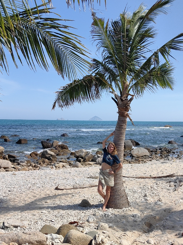 A woman stands leaning against a palm tree on a beach. The beach is sandy with scattered rocks and pebbles. Beyond the beach, there's a blue ocean with a small, hilly island visible in the distance. Palm leaves frame the upper part of the image. The woman is wearing a dark t-shirt and a patterned wrap skirt. The sky is a clear blue, indicating a sunny day.