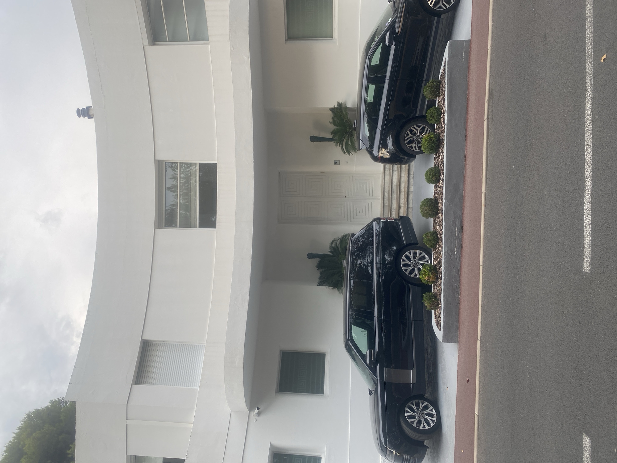 Two black SUVs are parked in front of a modern white building with curved architectural elements. The building has large windows and a prominent set of double doors with a geometric pattern. There are some potted plants and small shrubs placed in front of the vehicles and along the building's facade. The scene appears to be an exterior shot of a residential or commercial property.