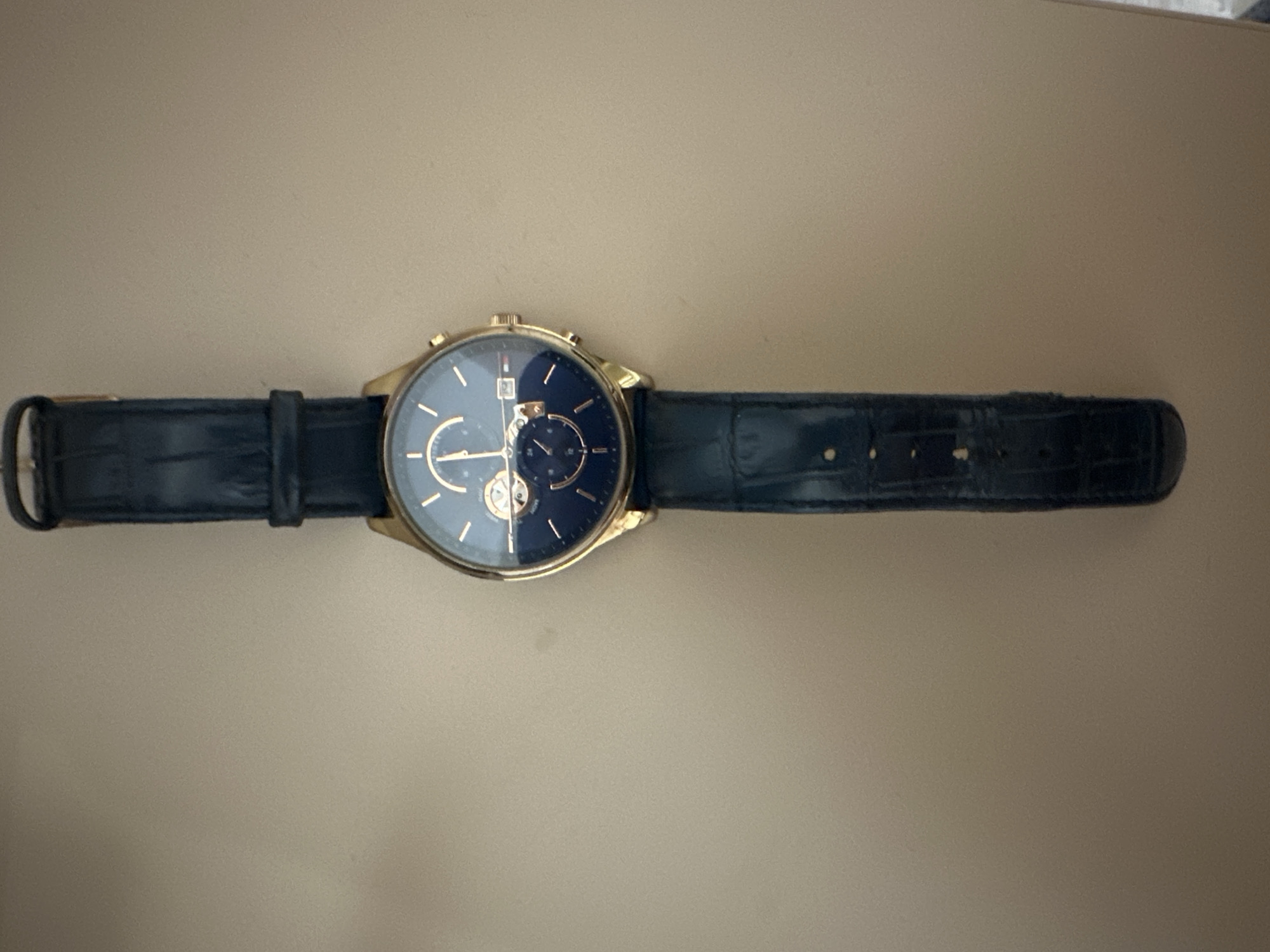 A gold-toned men's wristwatch with a dark blue dial and a dark blue leather strap is depicted laid flat on a neutral, light brown surface. The watch features gold-toned hour markers, hands, and two sub-dials. The upper sub-dial displays segments with text, possibly indicating days of the week or a chronograph function, with "GAU" and "MON" partially visible. The lower sub-dial is a 24-hour indicator marked "12" and "24". A date window at the 3 o'clock position shows the numeral "2". The brand name "TOMMY HILFIGER" is faintly visible below the 12 o'clock position. The watch case has two pushers flanking a crown on the right side. The leather strap exhibits an embossed crocodile or alligator pattern and includes a gold-toned buckle. The object is centered vertically, with the 12 o'clock position oriented towards the top of the frame. The background surface is uniform, indicating an indoor setting. The location is not discernible from the image.