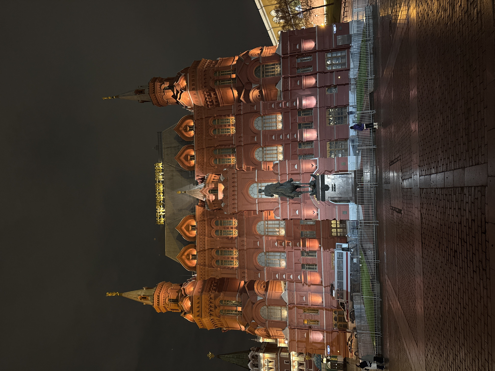 The image shows the State Historical Museum in Moscow, Russia, at night. The red brick building is lit up, revealing its architectural details, including towers, arched windows, and ornate decorations. A statue stands in front of the museum. The ground is brick-paved and reflects the building's lights. A fence separates the museum grounds from the paved area. People can be seen near the fence. The sky is dark.