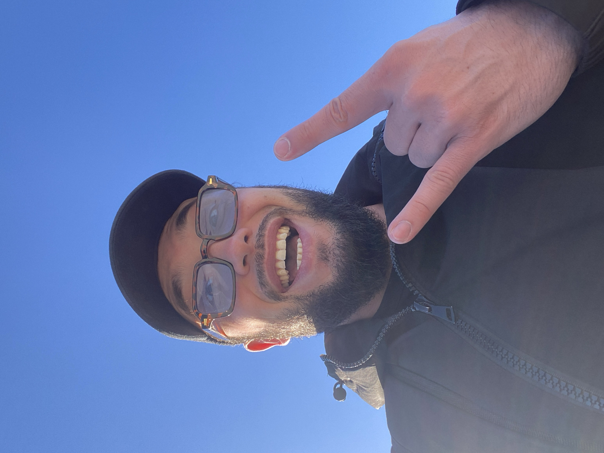 A man, wearing a black baseball cap, glasses with tortoise-shell frames, and a black jacket, is captured in a selfie against a clear blue sky. He has a beard and is smiling broadly, with his mouth open, revealing his teeth. His right hand is raised, forming the "rock on" or "horns" gesture. The lower part of the image shows the zipper of his jacket. The image appears to have been taken outdoors, with the bright sunlight casting a glare on the left side of the frame. The background is uniformly blue, indicating an unobstructed sky. There is no discernible urban or natural environment beyond the man and the sky. The location in Koskolovo, Russia is not indicated by any visual cues in the image itself.